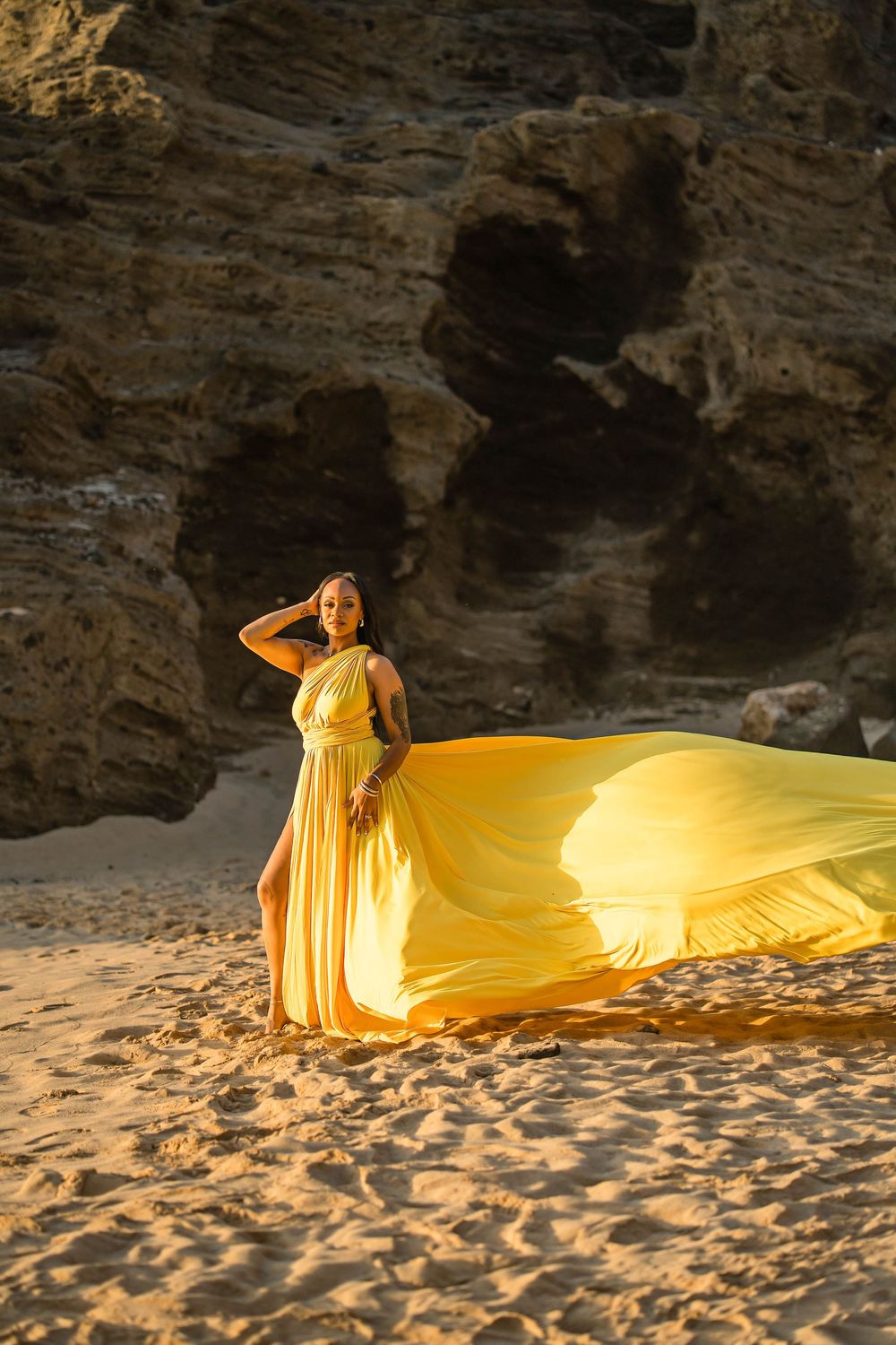 A flowing yellow dress creates a dramatic trail along a sandy beach against rocky cliffs at sunset.