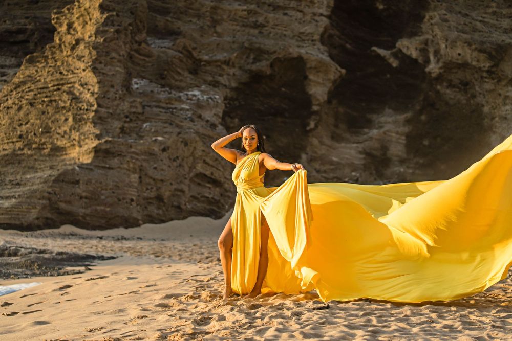 Elegant yellow gown flows in the wind against dramatic rocky beach backdrop during sunset photo shoot.