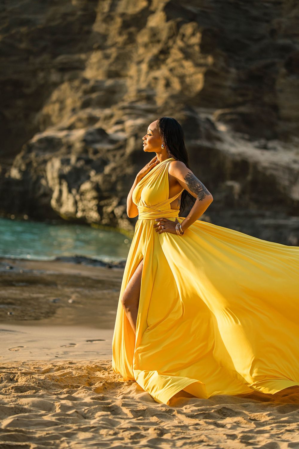 A flowing yellow dress catches the wind on a beach at sunset with dramatic rocky cliffs in the background.