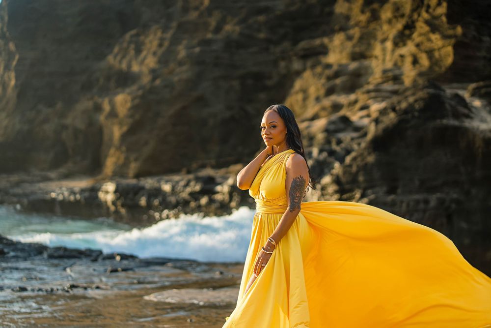 A woman in a flowing yellow dress stands gracefully against a rocky coastal backdrop as waves crash behind her.