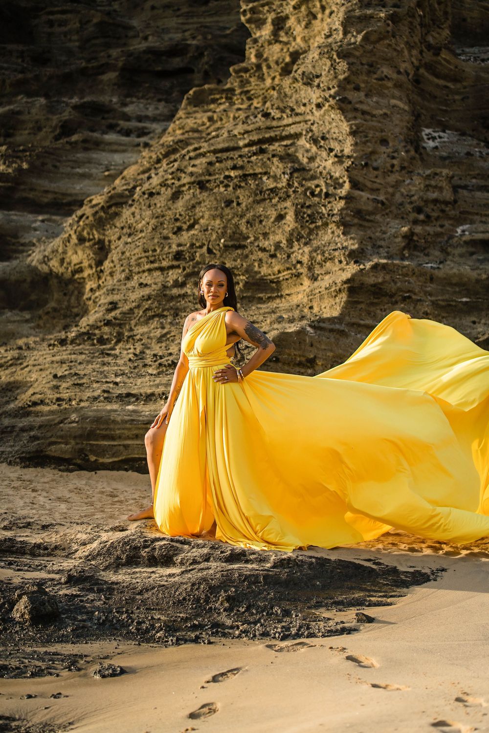 A flowing yellow dress billows dramatically against rocky coastal cliffs and sandy beach during a sunset photoshoot.
