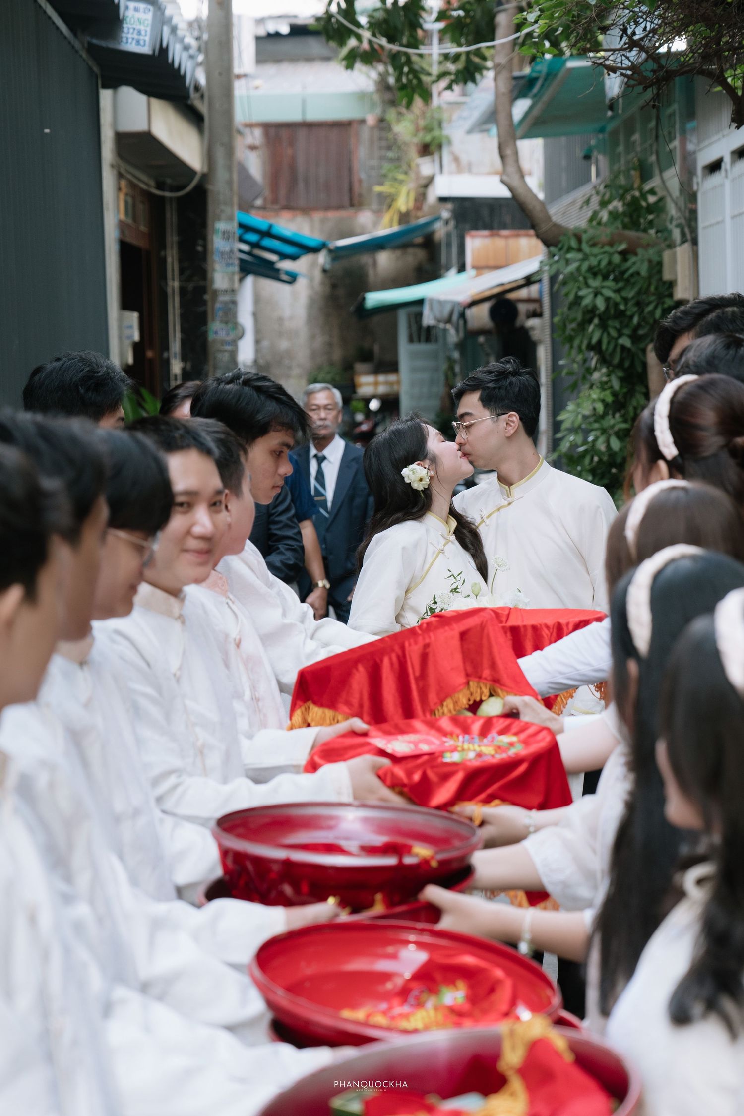 Vietnamese bride and groom share a kiss during traditional wedding procession in a narrow alley.