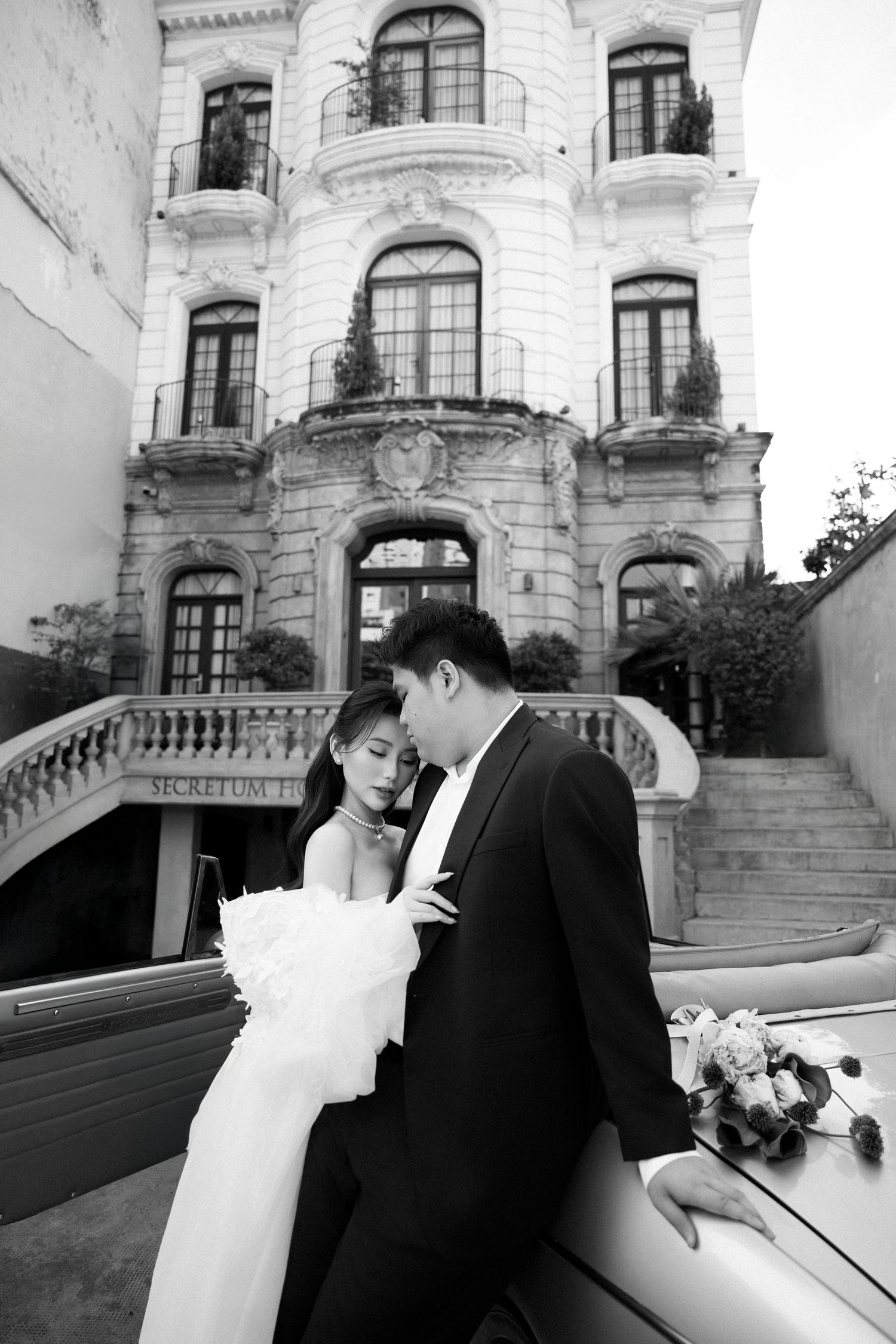 Black and white photo of bride and groom embracing beside vintage car at Secretum Hotel.