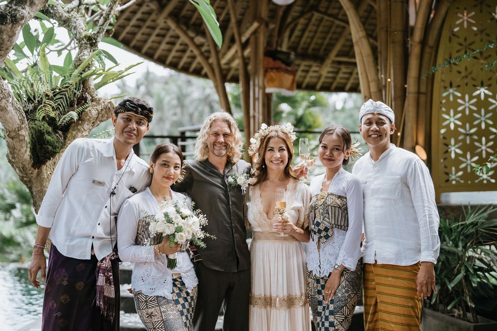 Wedding guests gather under a thatched roof pavilion decorated with flowers and lanterns.
