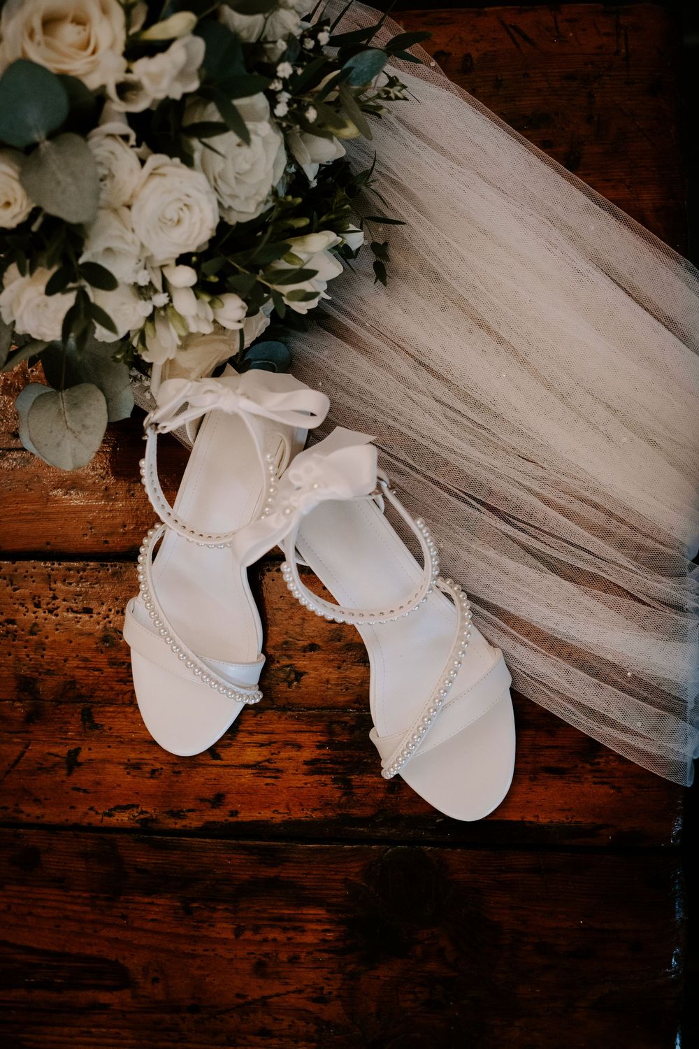 Elegant white bridal sandals with pearl straps next to roses and veil on wooden surface.