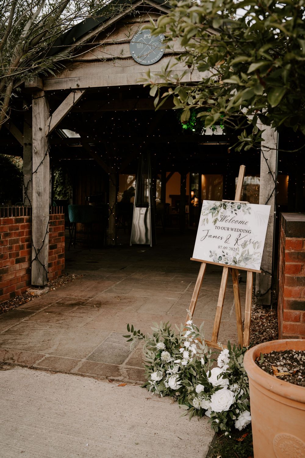 Elegant wedding welcome sign display with white floral arrangements and wooden easels at venue entrance.