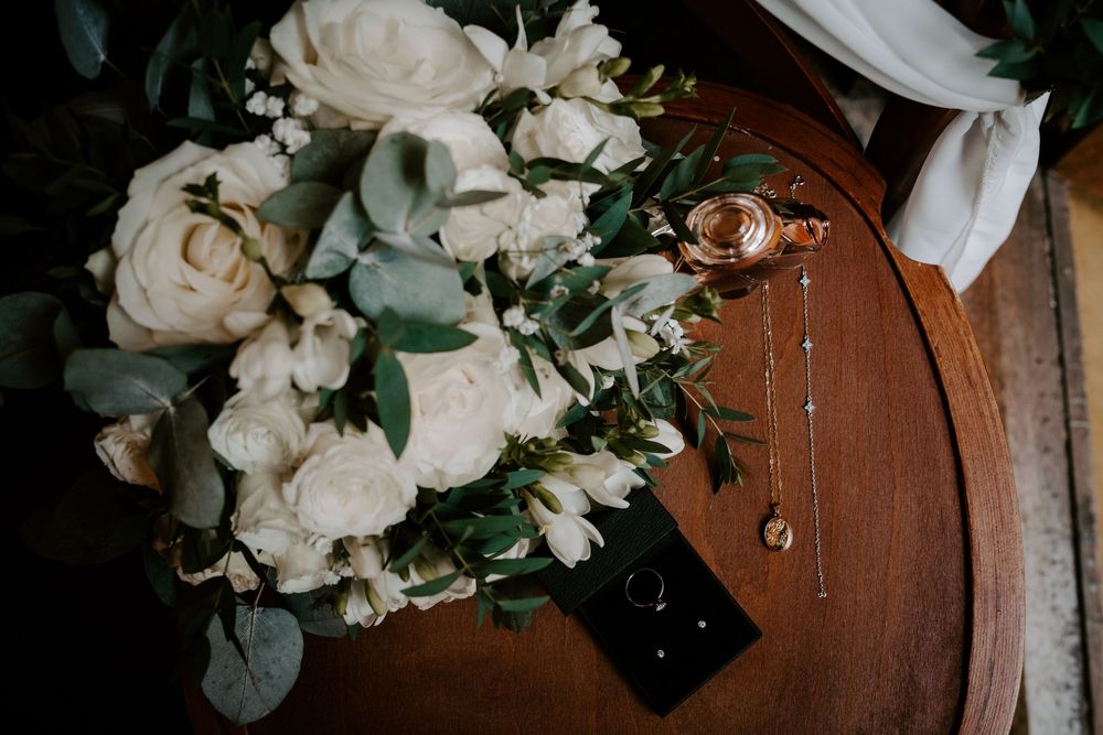 White floral wedding bouquet with greenery and wedding rings displayed on rustic wood surface.