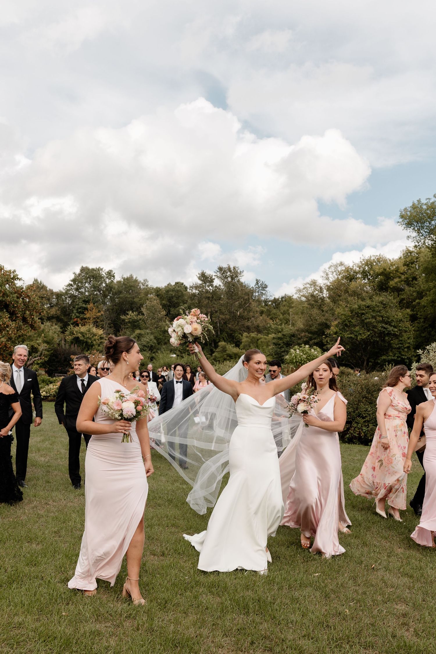 A wedding party in pink dresses and formal attire dances and celebrates joyfully in a grassy outdoor field.