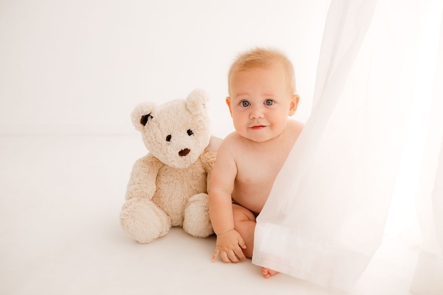 Baby in white outfit lying next to plush teddy bear on white background.