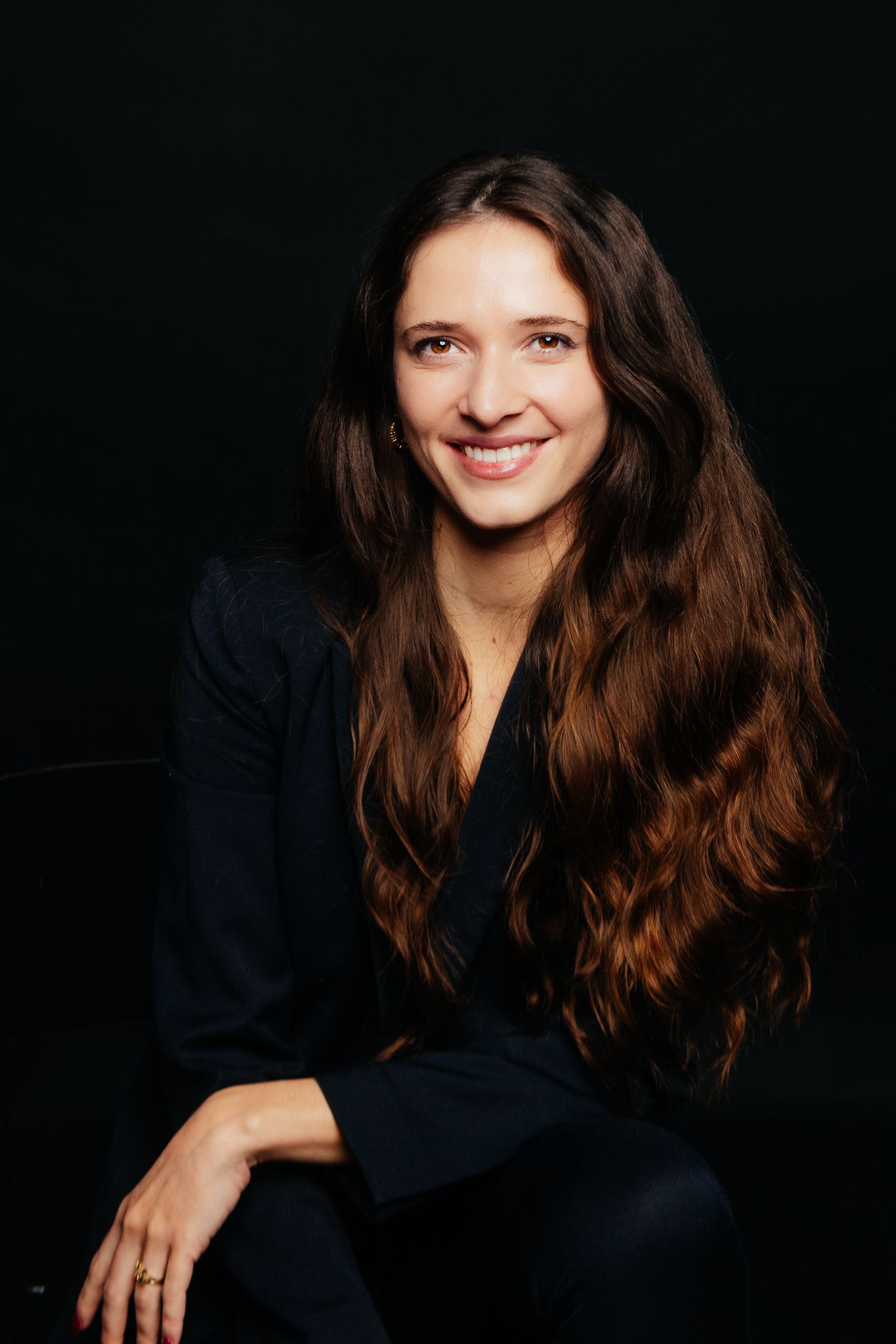 Young woman with long wavy brown hair smiling in black outfit against dark background.