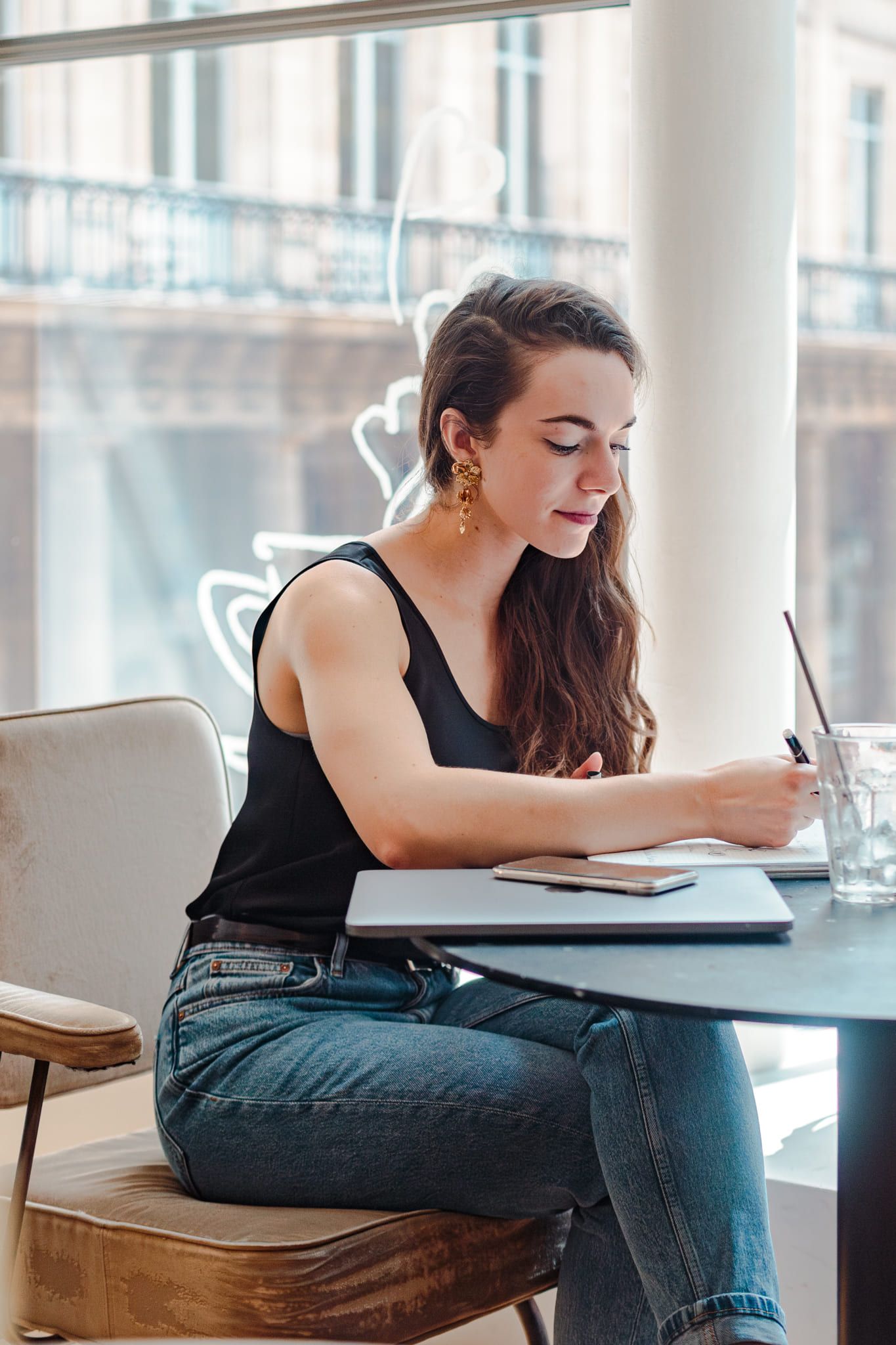 Young woman with earrings writing in notebook at cafe table by bright window.