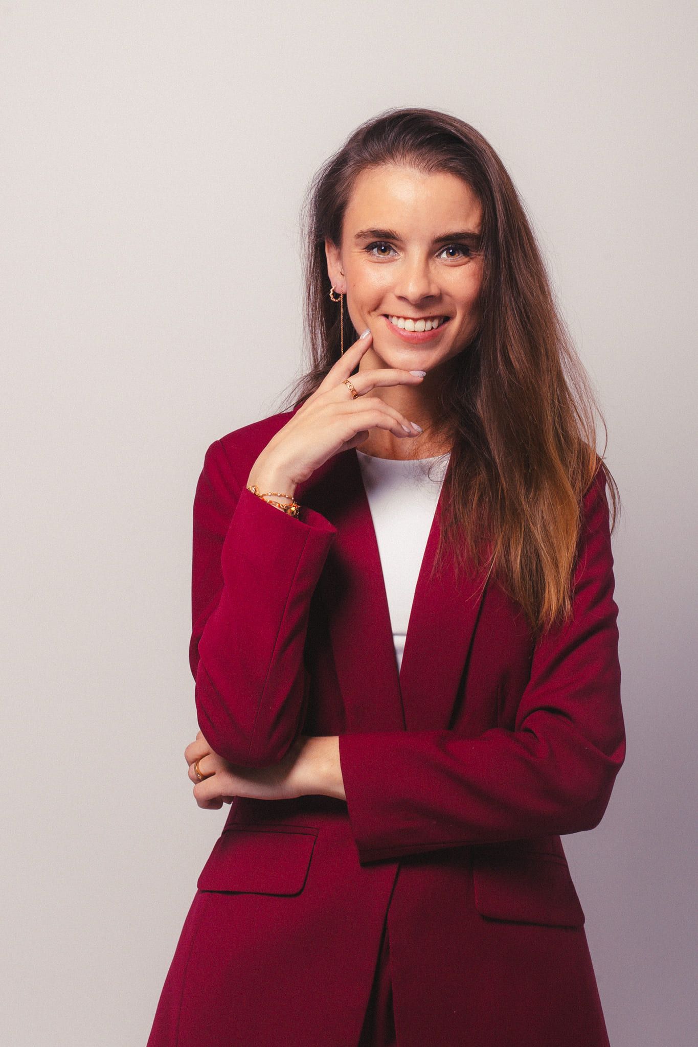 Smiling woman in red blazer posing with hand on chin against gray background.