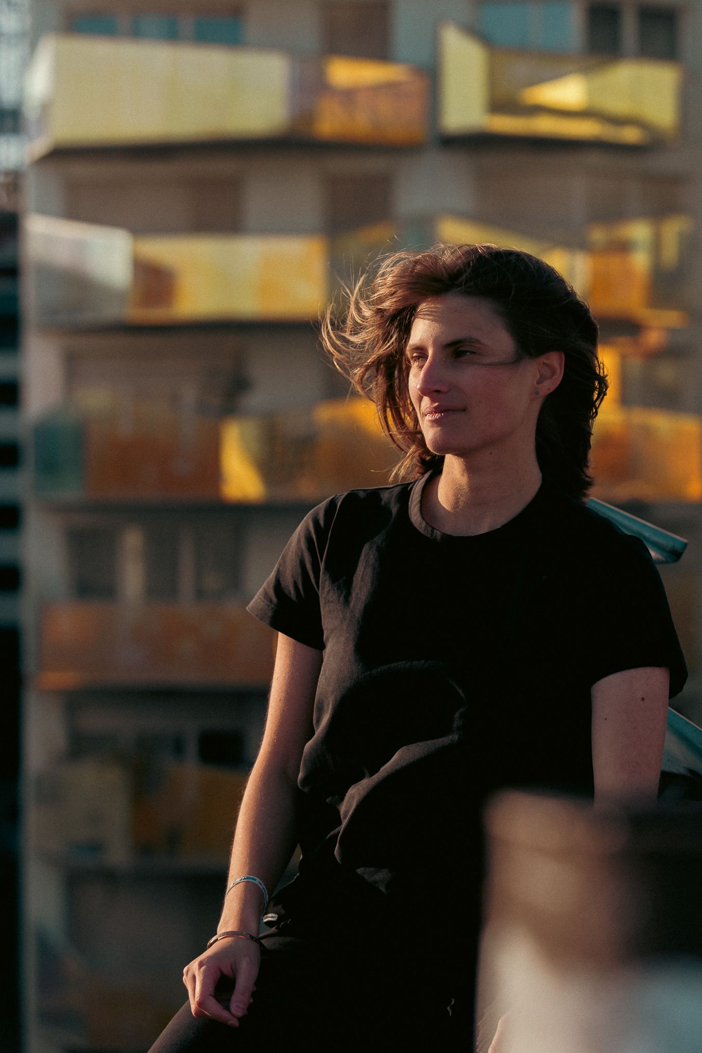 Young woman in a black t-shirt with wind-blown hair standing on an urban rooftop at sunset.