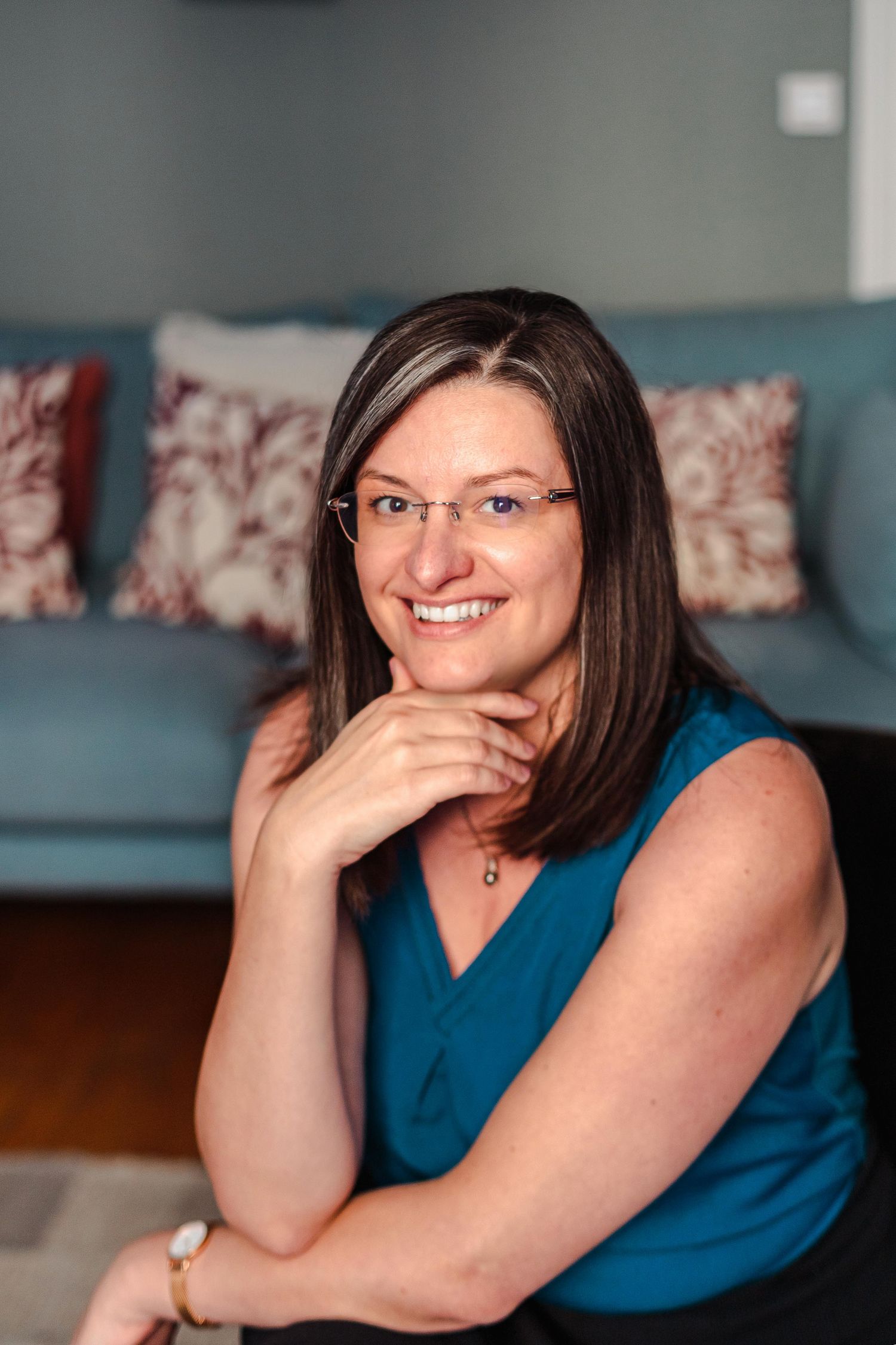 Smiling woman with glasses and dark hair in teal top seated in bright living room.