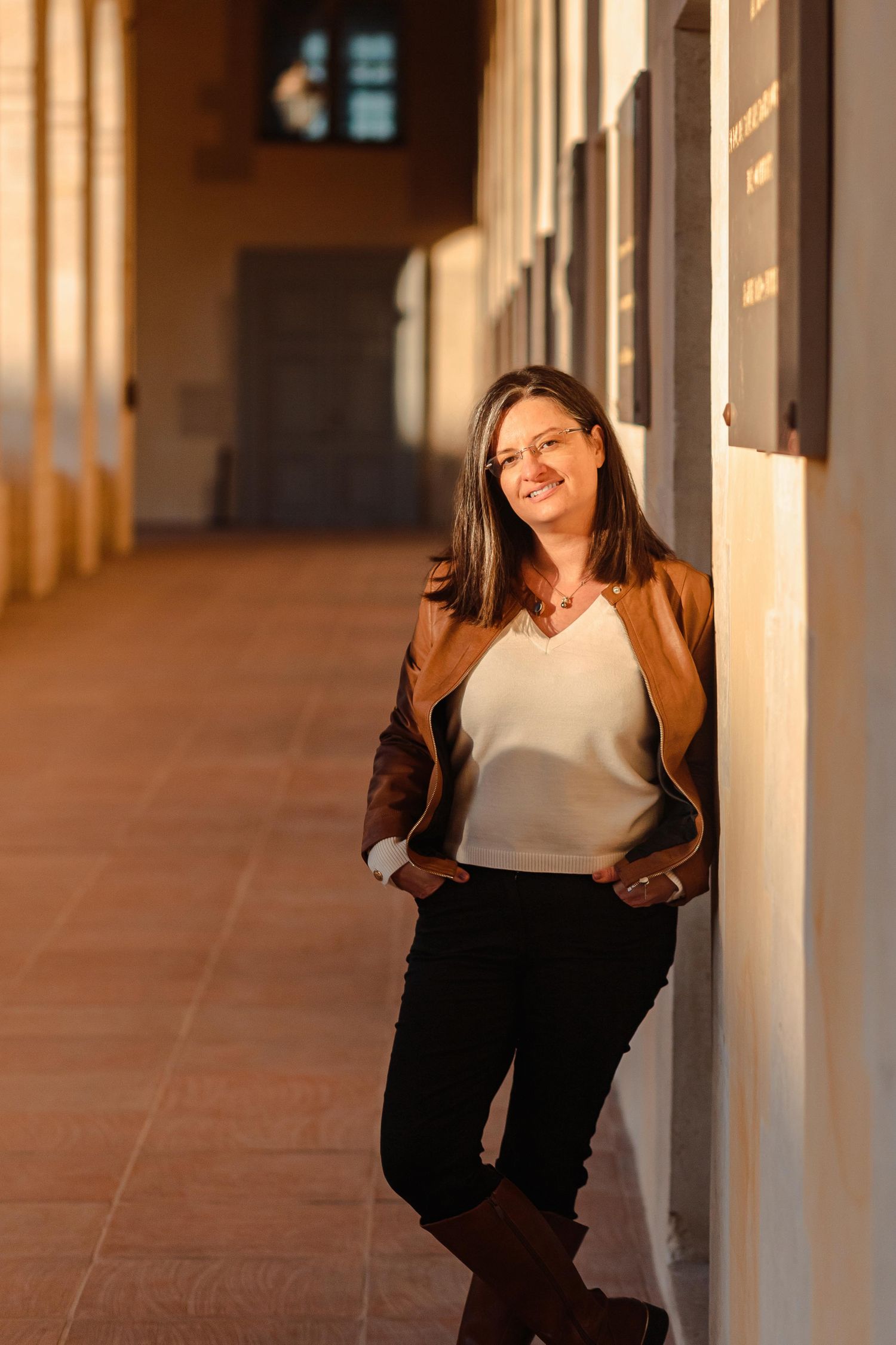Woman in brown jacket leaning against wall in sunlit corridor with warm golden light.