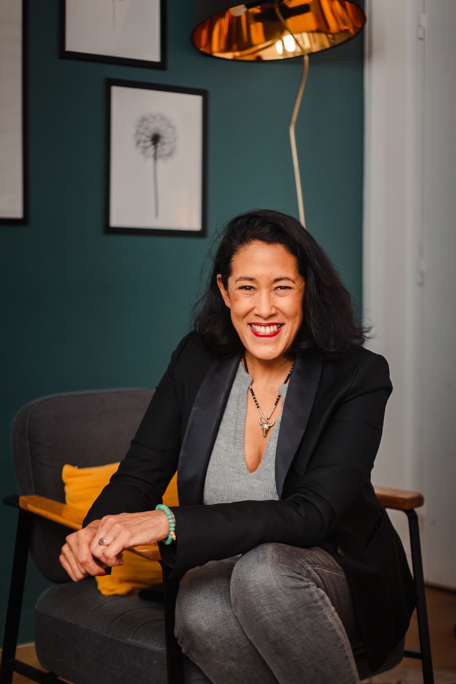 Smiling woman in black blazer seated in modern interior with teal wall and lamp.