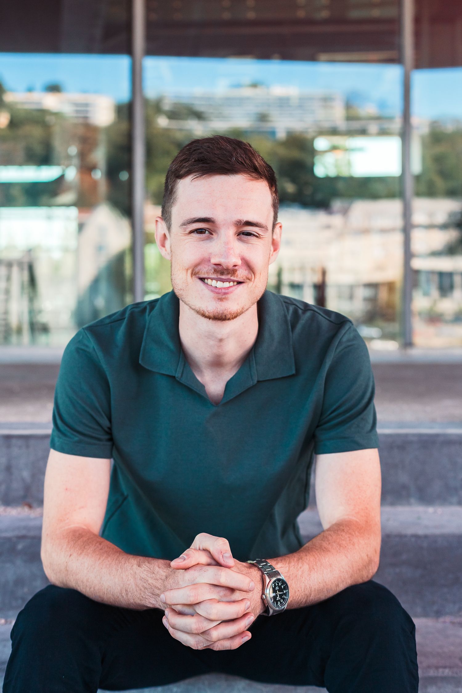 Young smiling man in green polo shirt sitting on steps outside modern building.