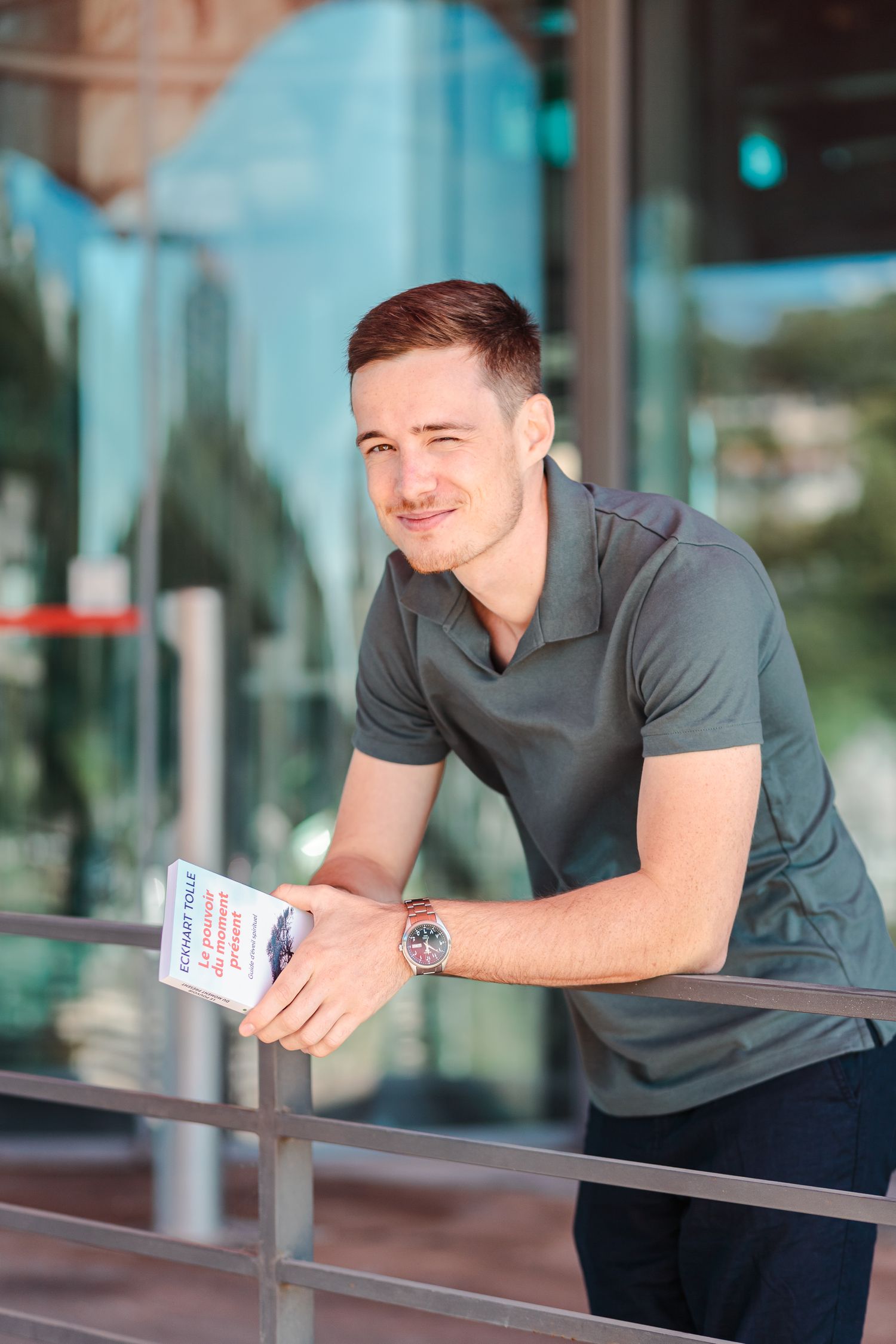 Young man in gray polo shirt leaning on railing holding book outside glass building.