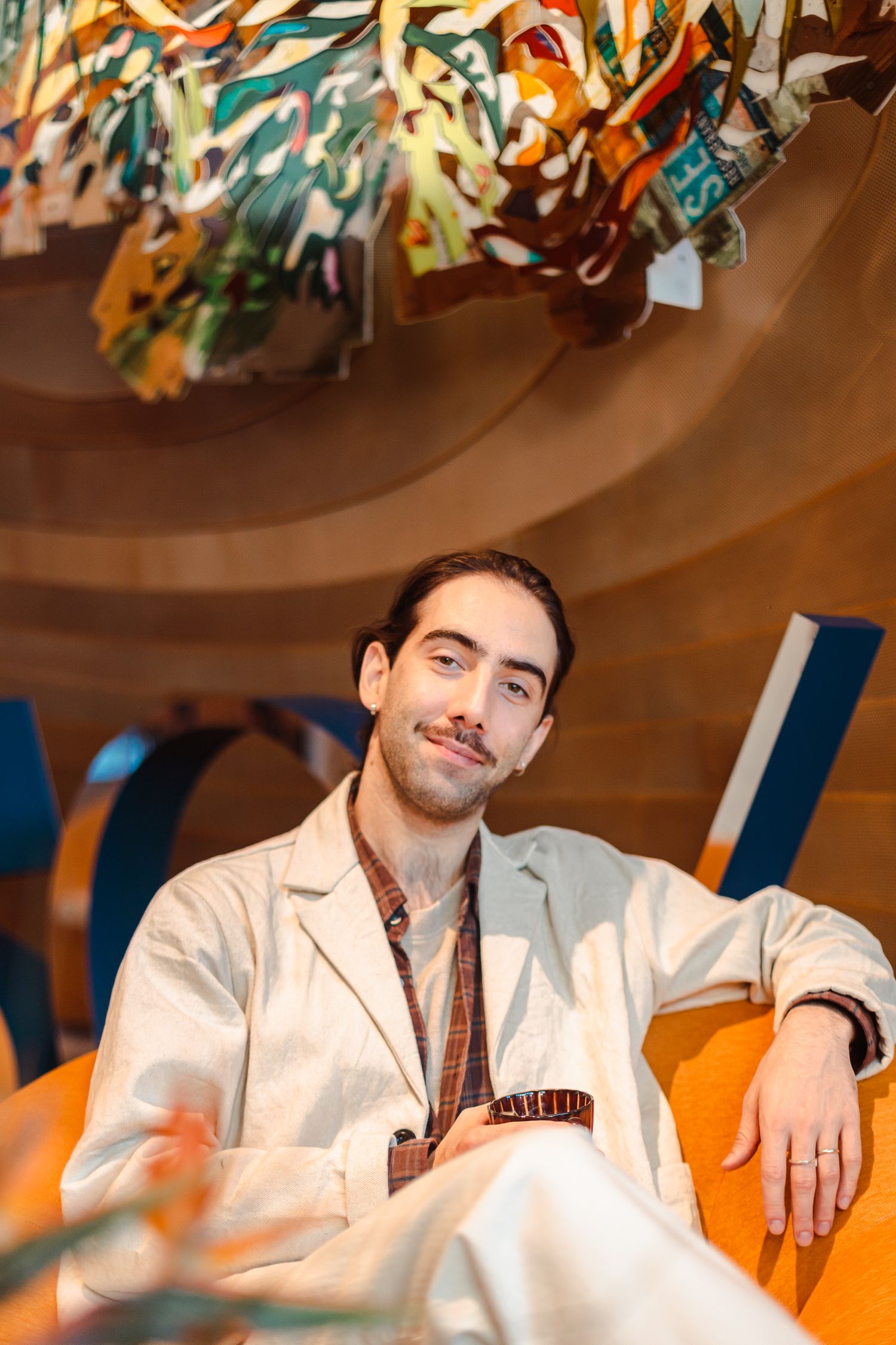 Young man in a white jacket sitting casually under a colorful art installation indoors.