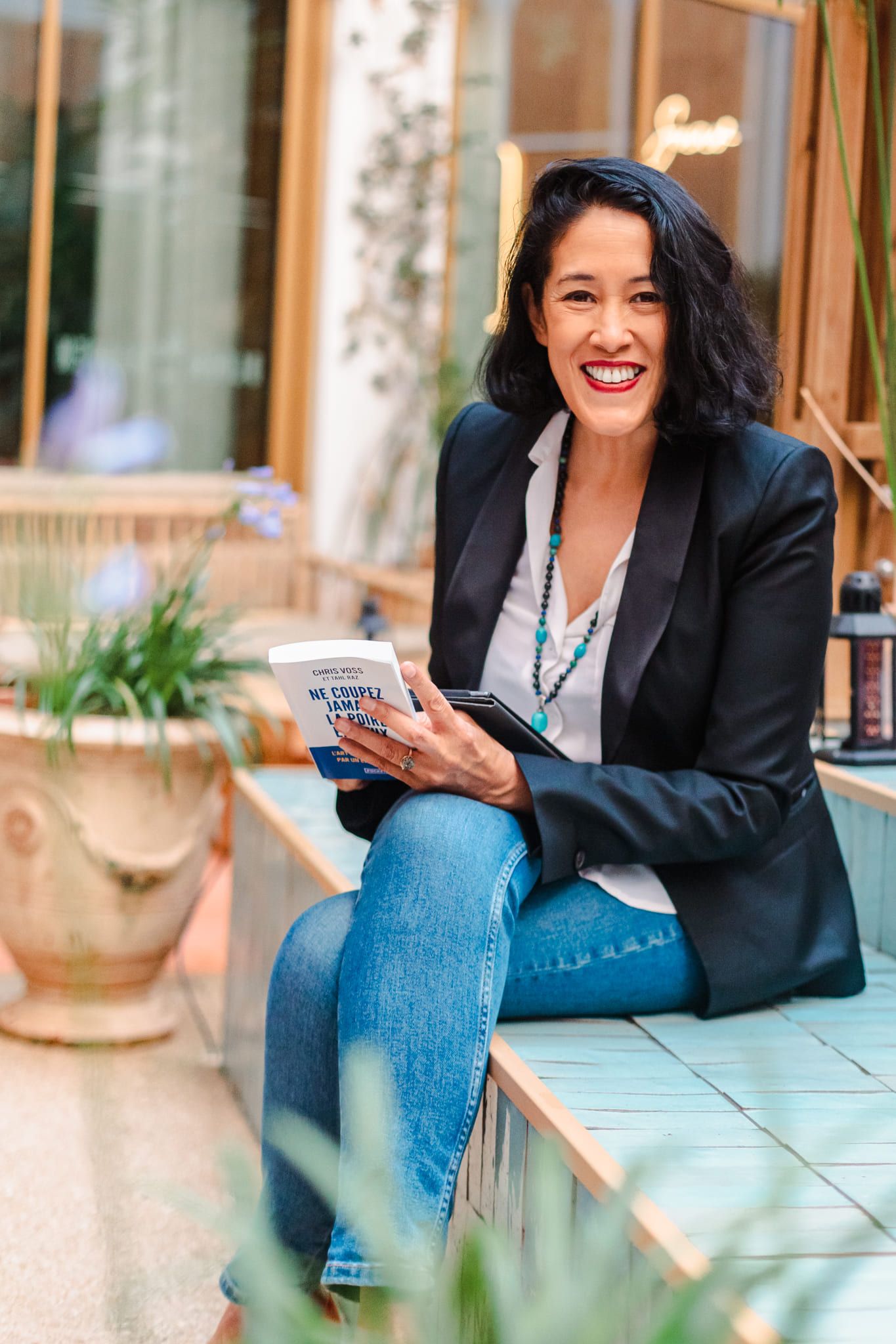 Smiling woman in black blazer holding book while sitting outdoors in courtyard.