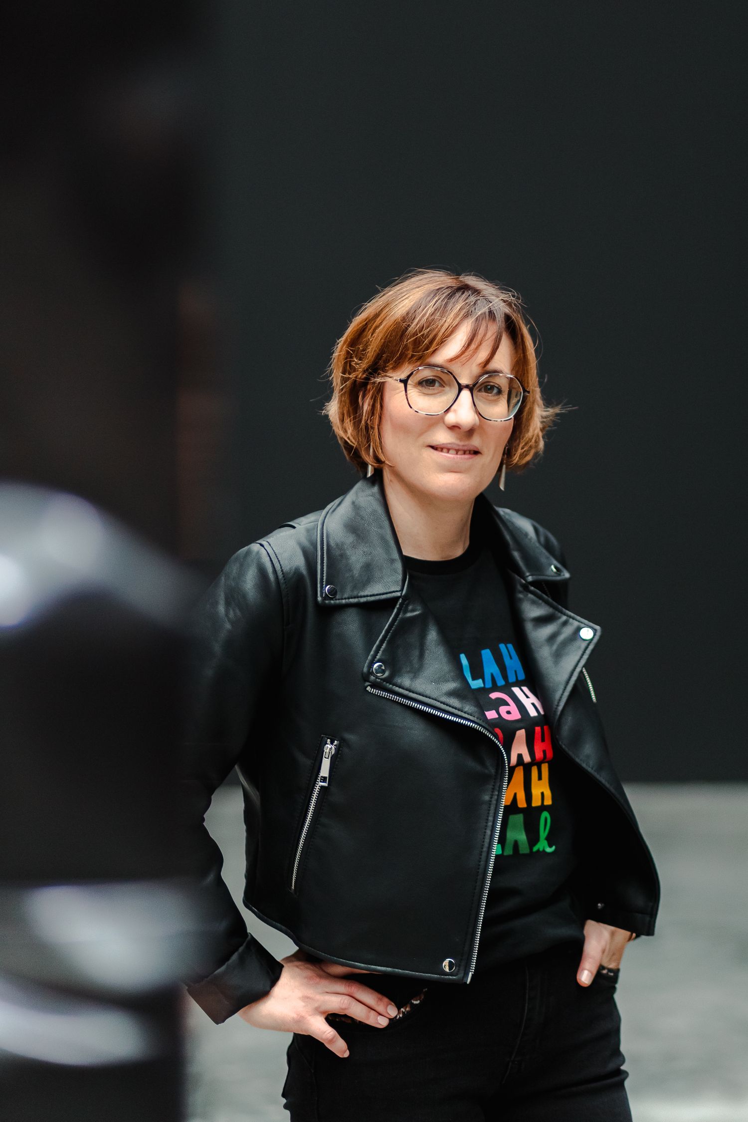 Woman in leather jacket and colorful graphic tee posing outdoors with hands on hips.