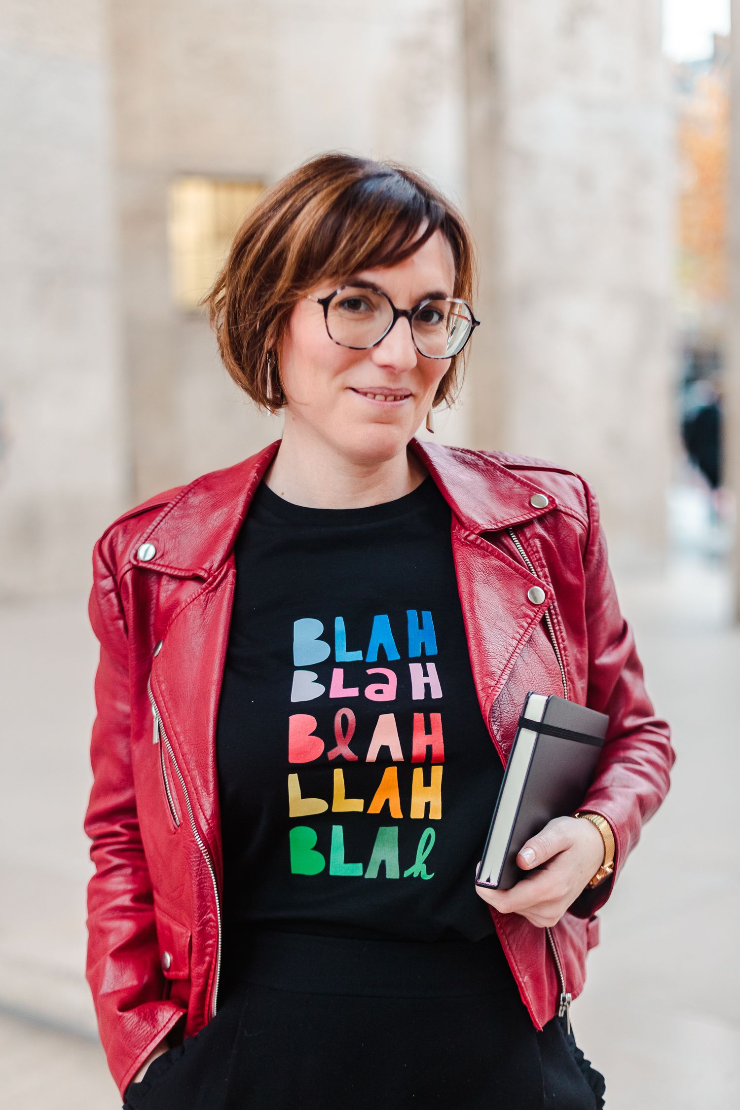 Woman in red leather jacket and Blah Blah t-shirt holding notebooks outdoors.