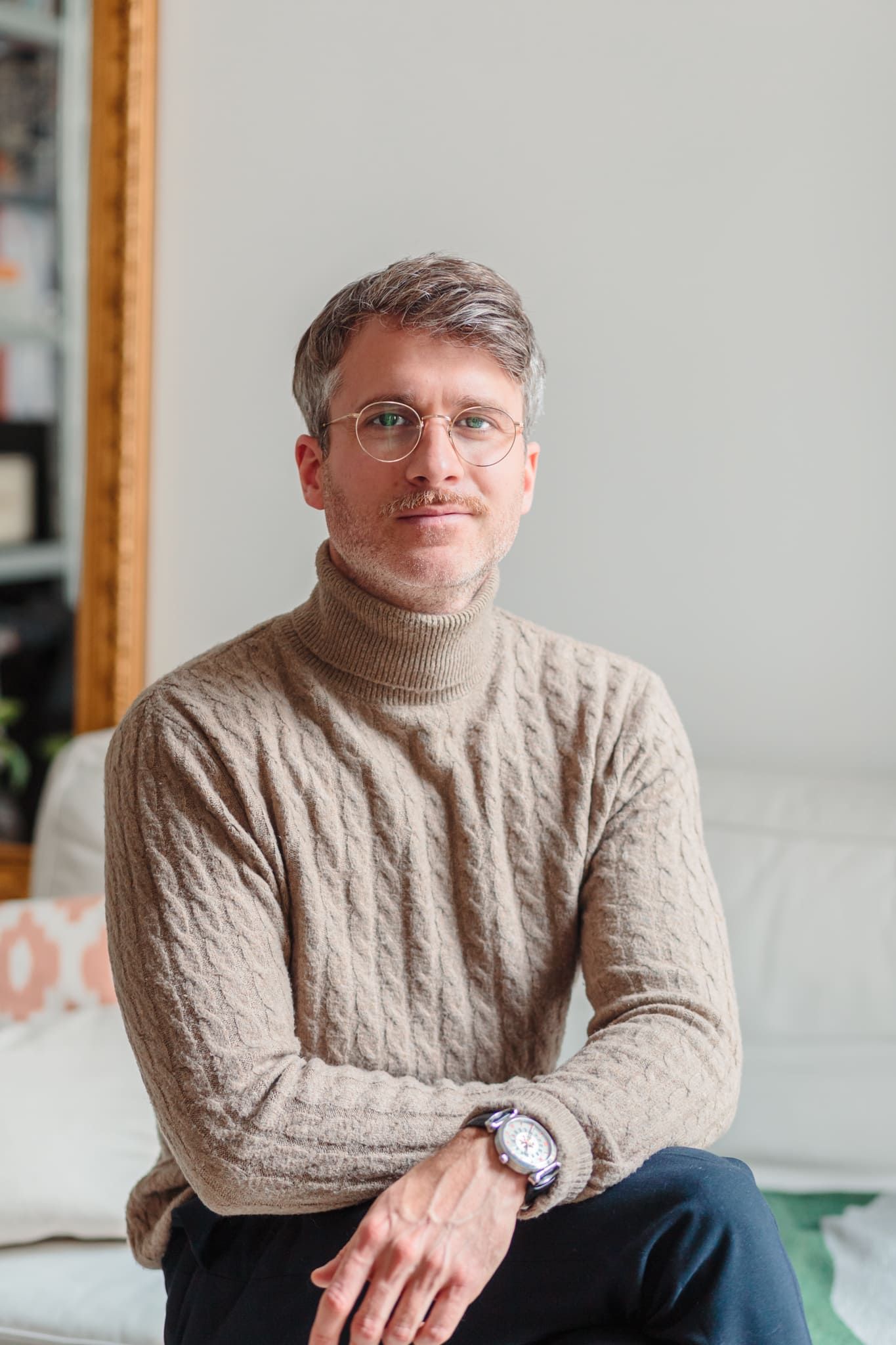 Man with glasses in beige turtleneck sweater sitting relaxed in bright home interior.