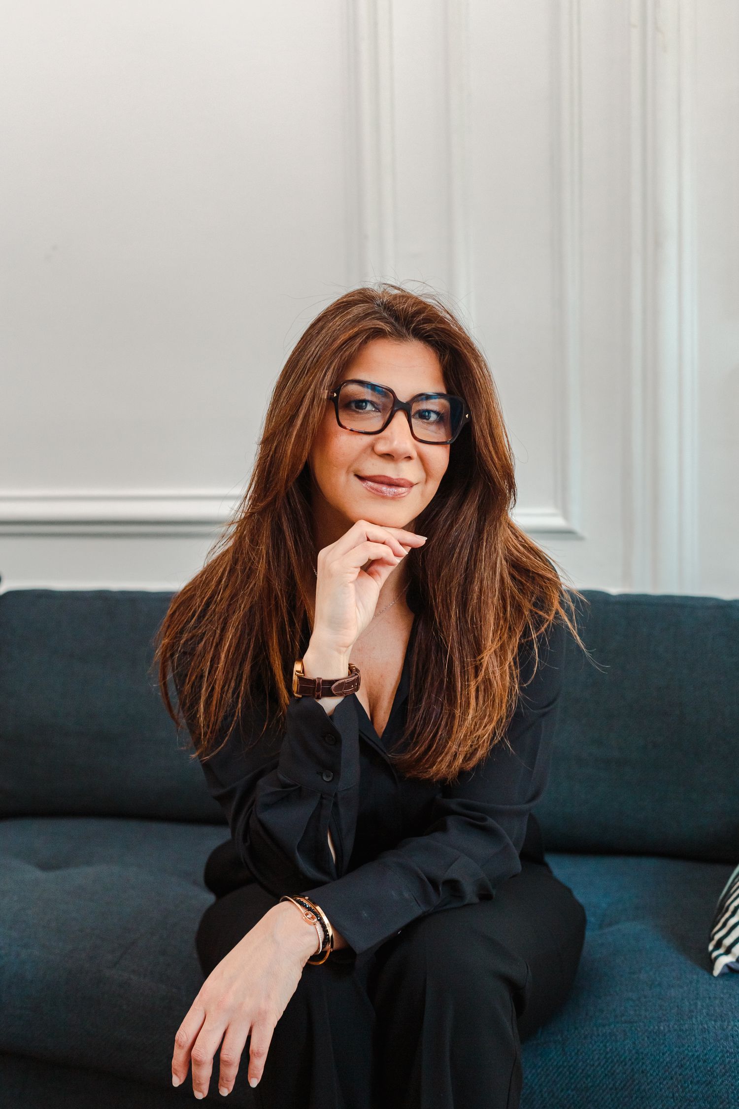 Elegant woman with glasses and auburn hair seated on blue sofa in white interior.