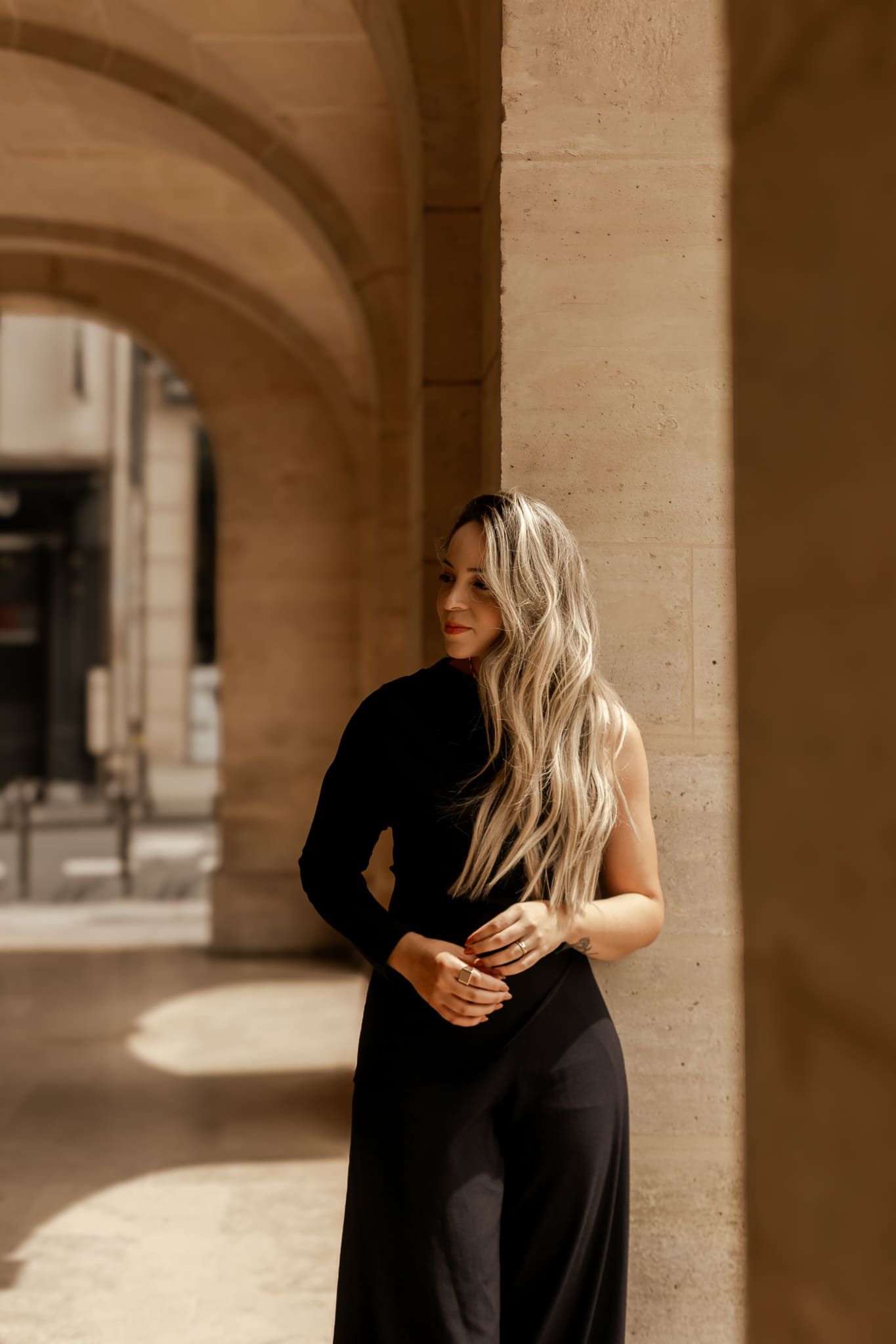 Elegant blonde woman in a black dress leaning against stone columns in a classical arcade.