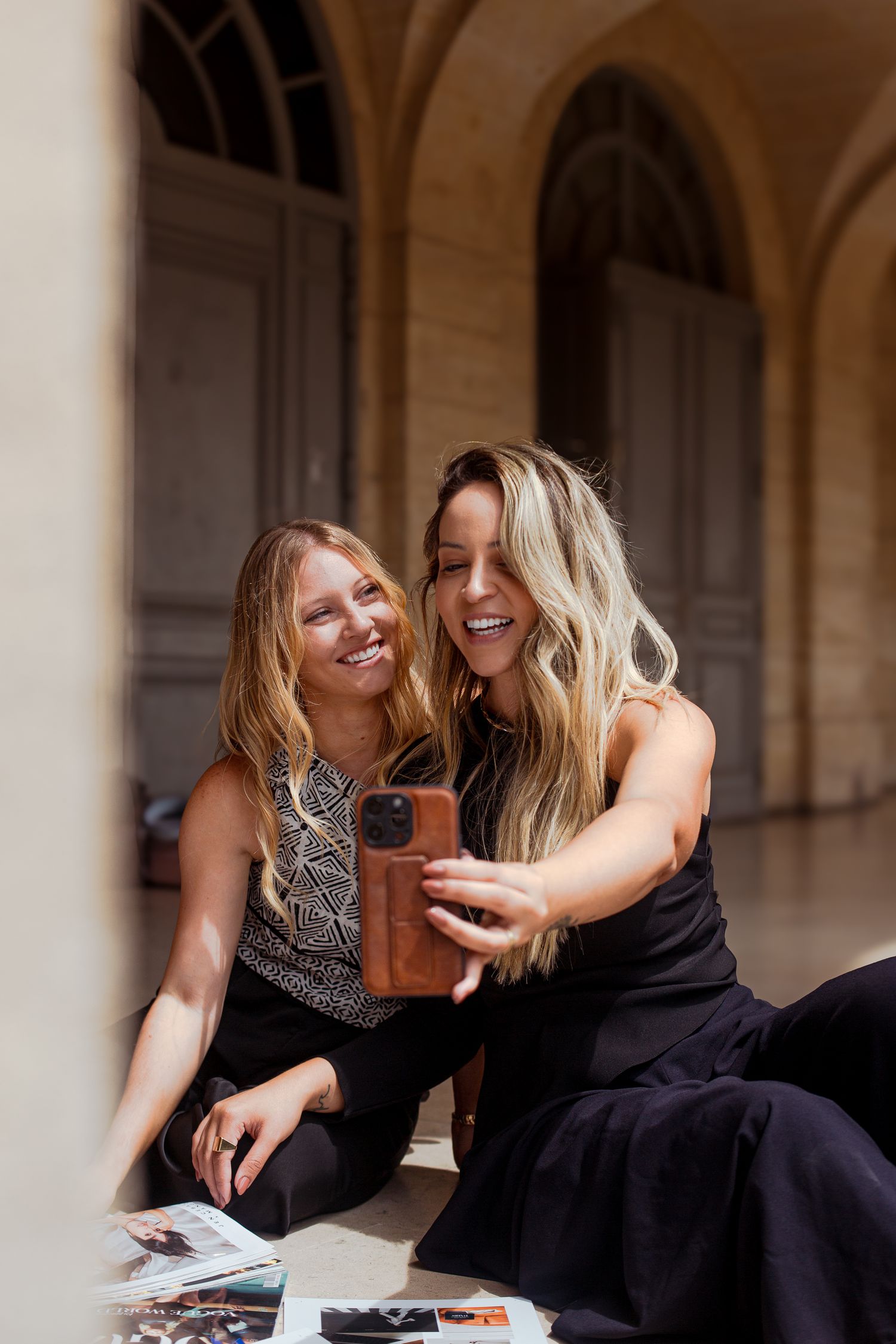 Two blonde women laughing while taking a selfie outdoors in elegant archway setting.
