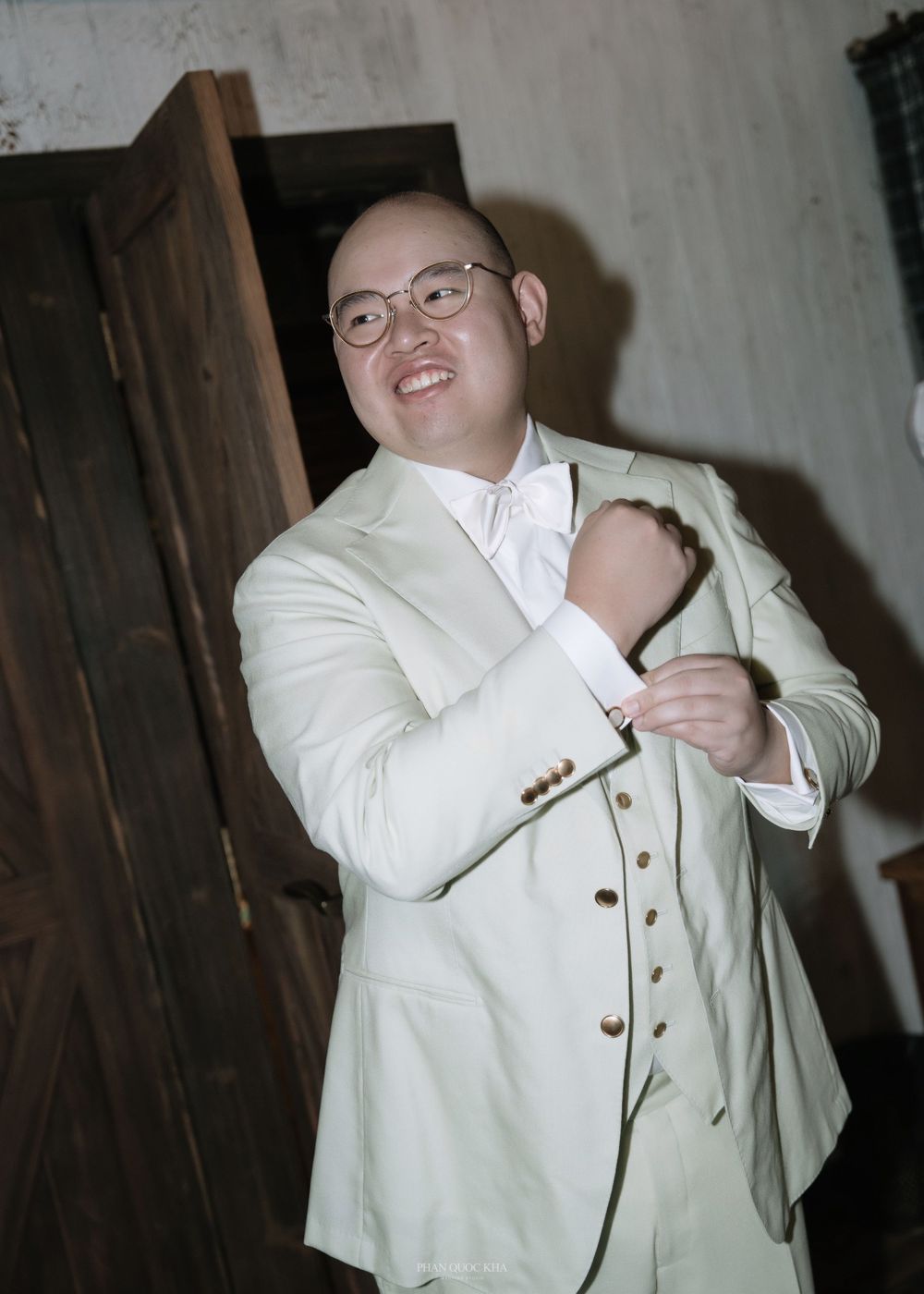 Smiling bald groom in a light green suit adjusting his cufflinks while getting ready for his wedding ceremony.