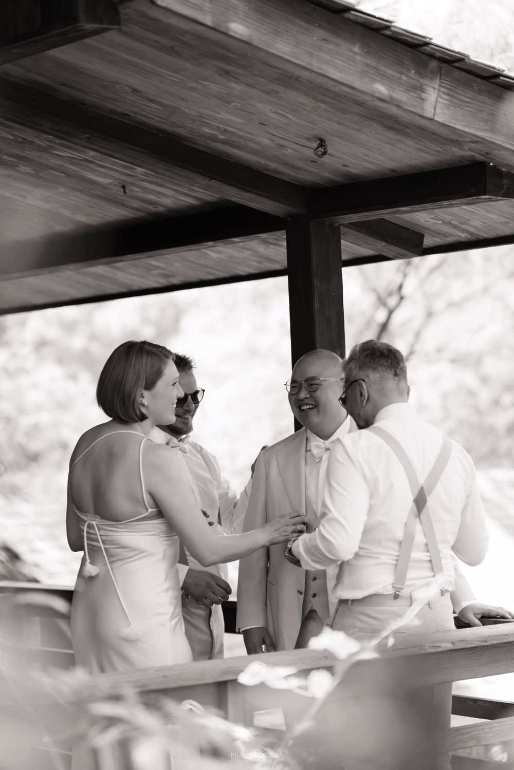 Black and white photo of groom laughing with groomsmen and guests under a wooden pavilion before the ceremony.