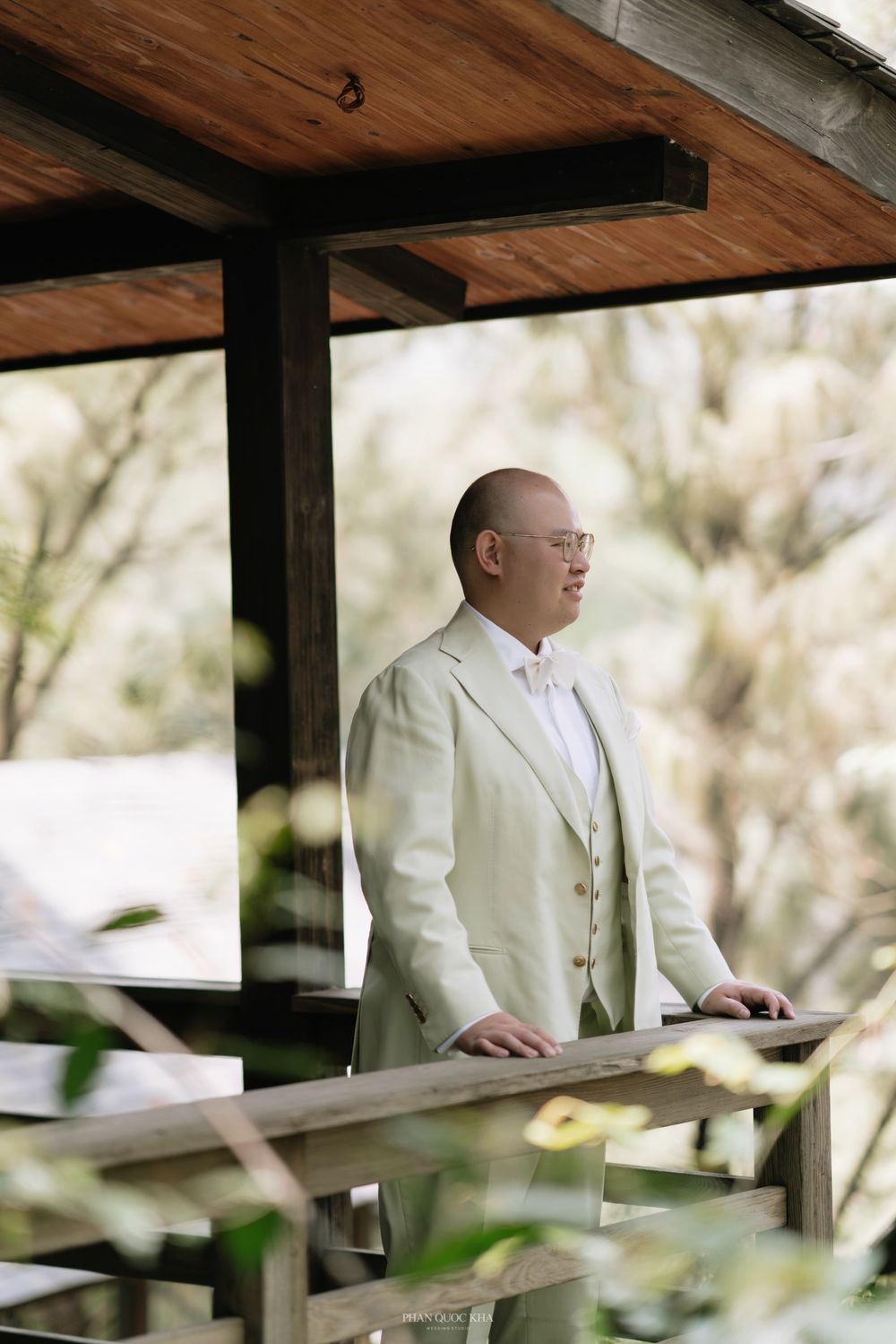 Groom in ivory suit stands on wooden deck surrounded by lush greenery at outdoor wedding venue.