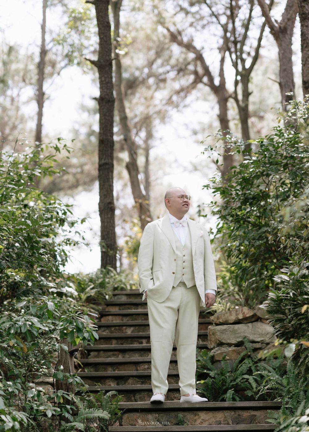 Groom in cream three-piece suit stands on stone steps surrounded by lush garden greenery.