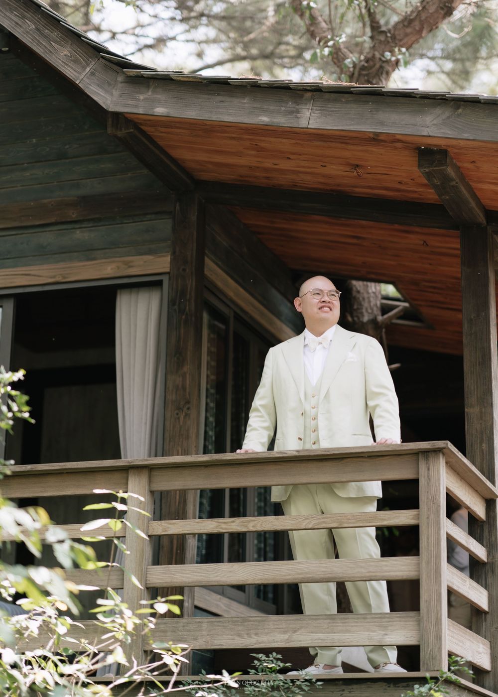 Smiling groom in ivory three-piece suit stands on rustic wooden cabin porch at wedding.