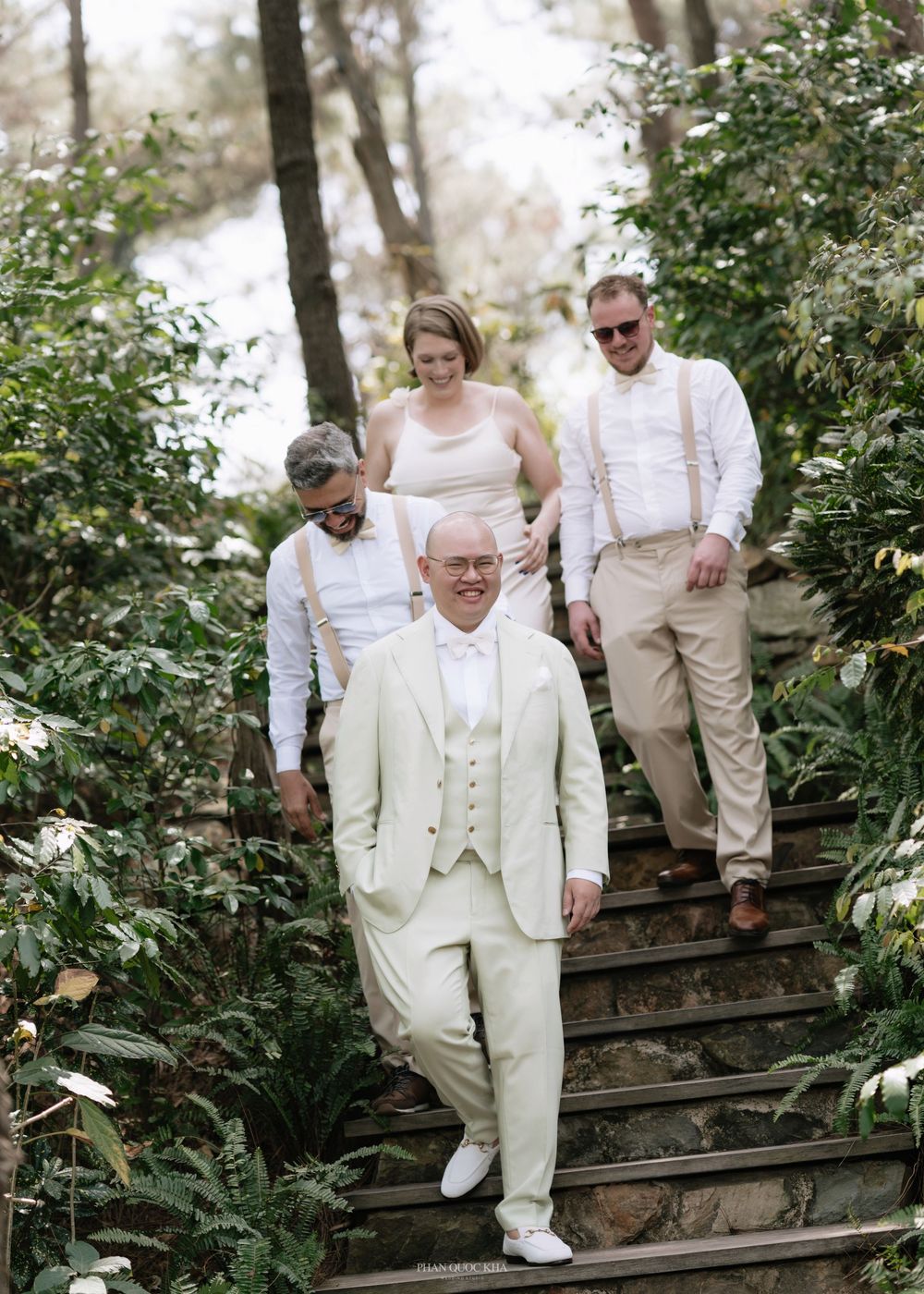Groom in ivory suit leads groomsmen and bridesmaid down stone garden steps at wedding.