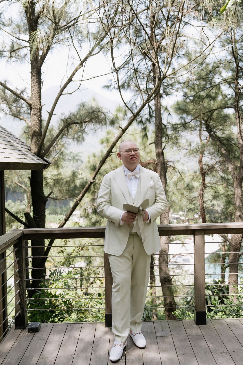 Groom in cream suit holds book on wooden deck surrounded by tall pine trees.