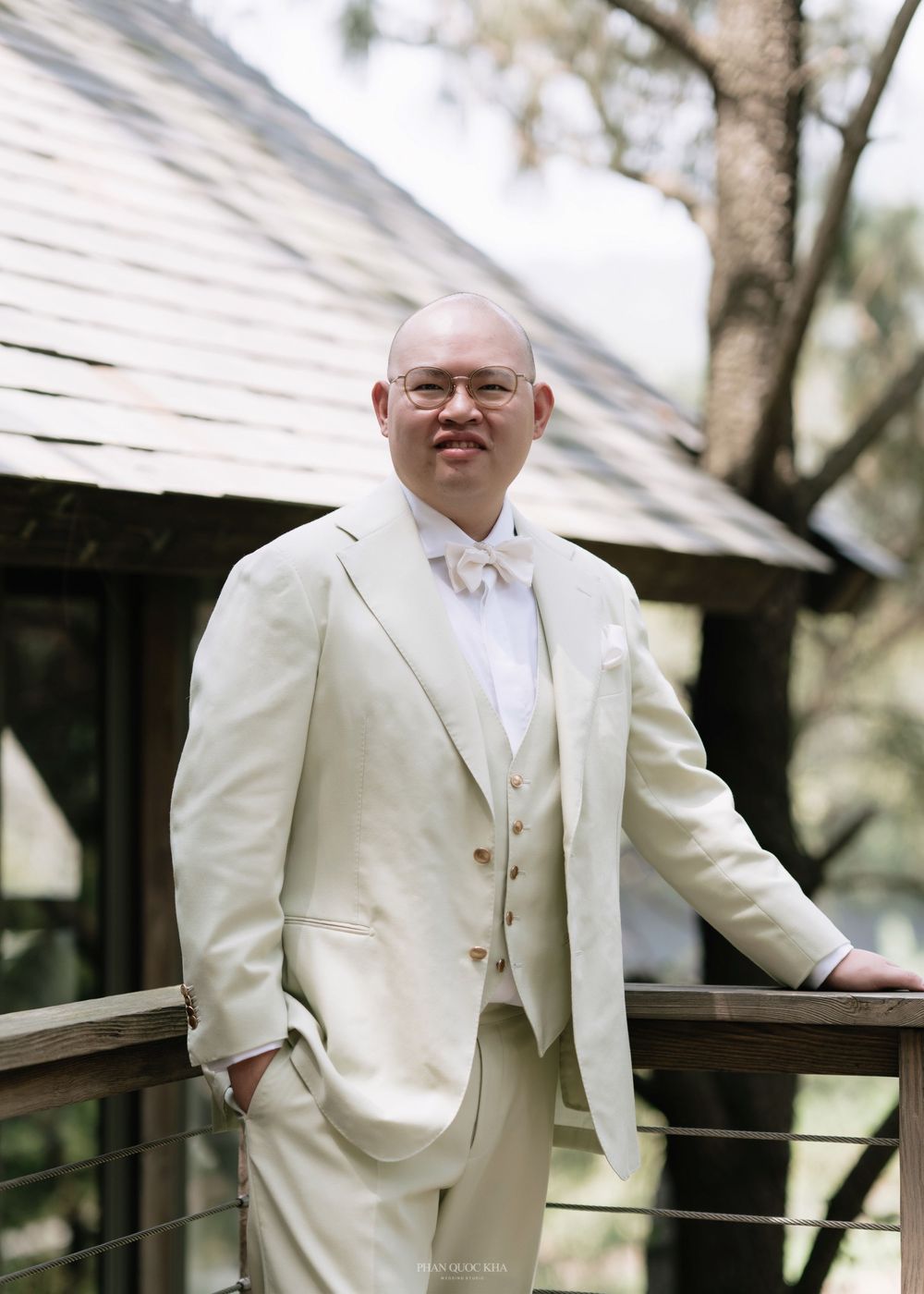 Groom in cream three-piece suit with bow tie poses by railing at outdoor wedding.