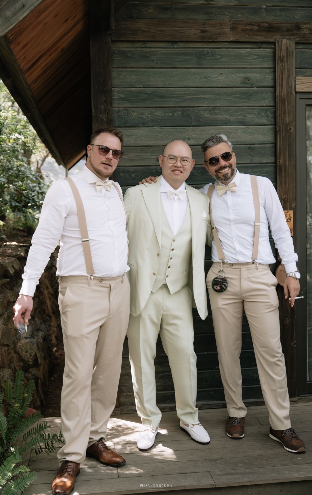Groom in ivory suit poses with two groomsmen in tan suspenders outside wooden venue.