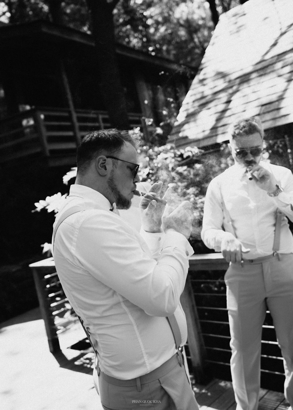 Black and white photo of groomsmen smoking cigars outdoors in white shirts and suspenders at a wedding.