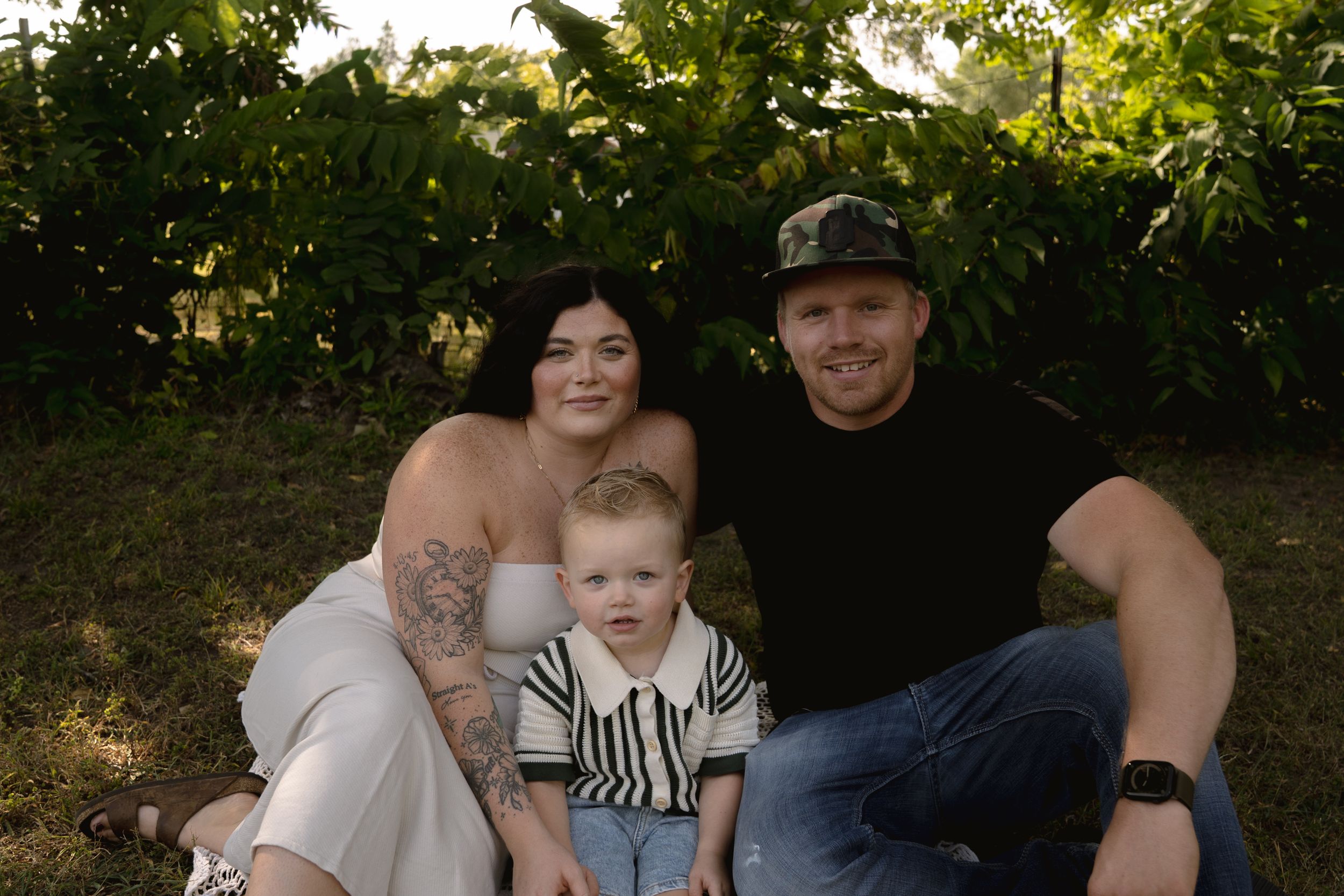 Family sitting together outdoors under green trees during a summer photoshoot.