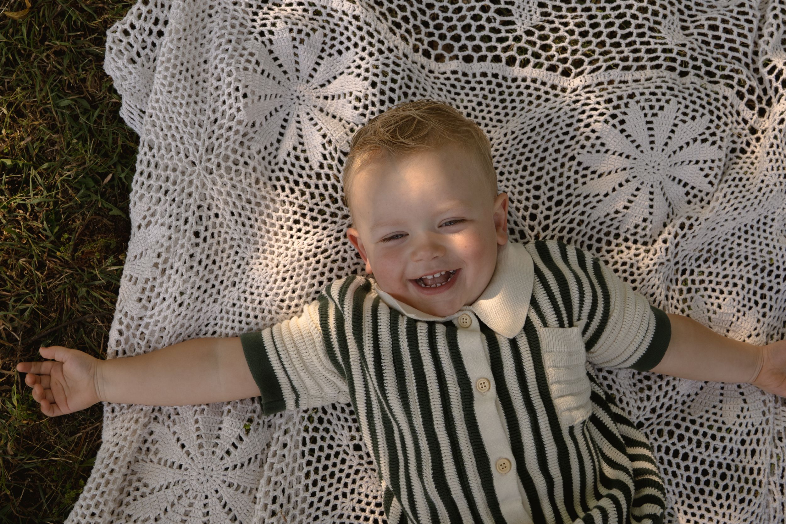 Child in striped outfit smiles while lying on intricate lace blanket outdoors.