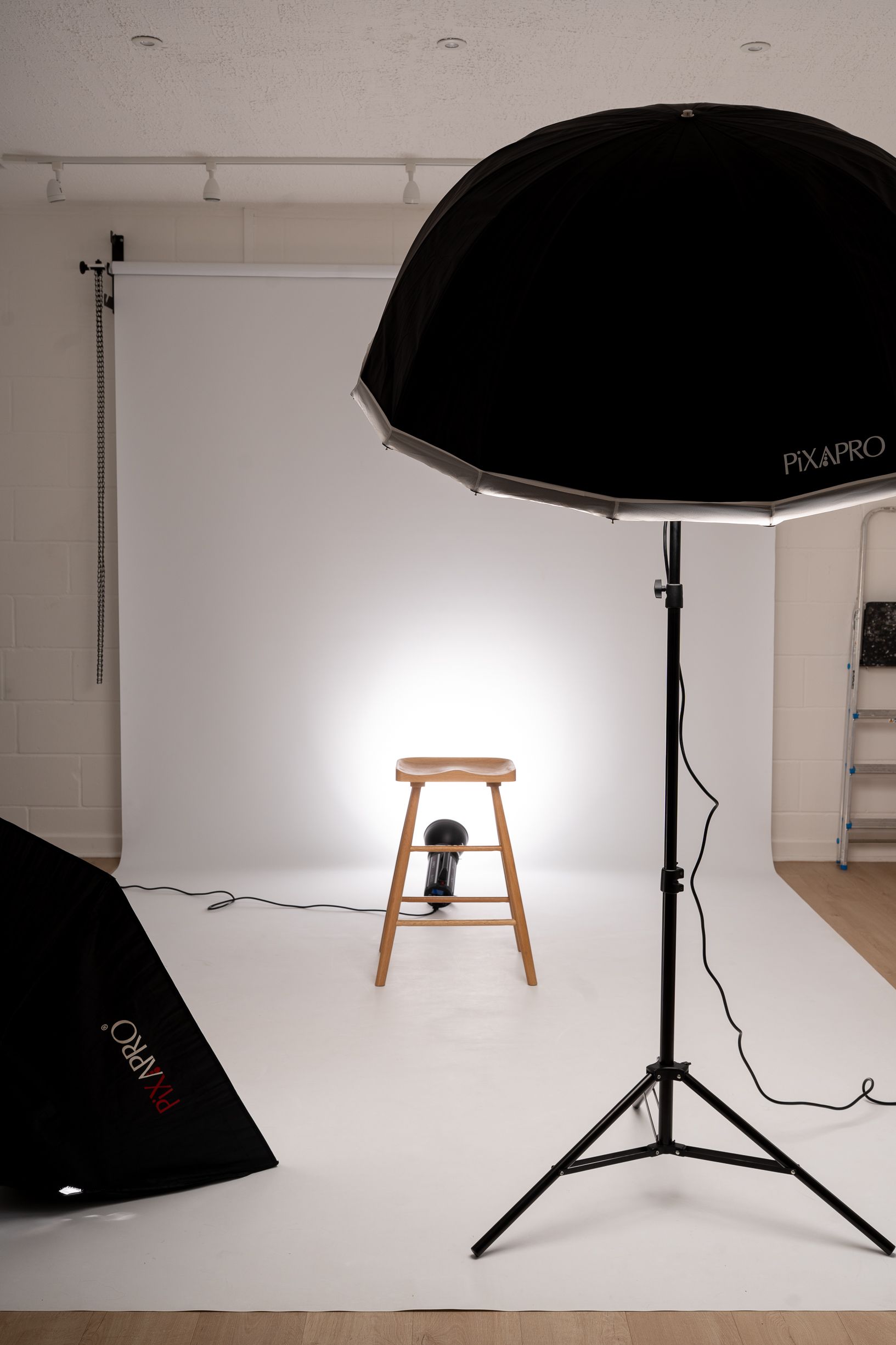 A photo studio set up with a white backdrop, a wooden stool and lighting equipment and umbrellas in Cambridge, UK