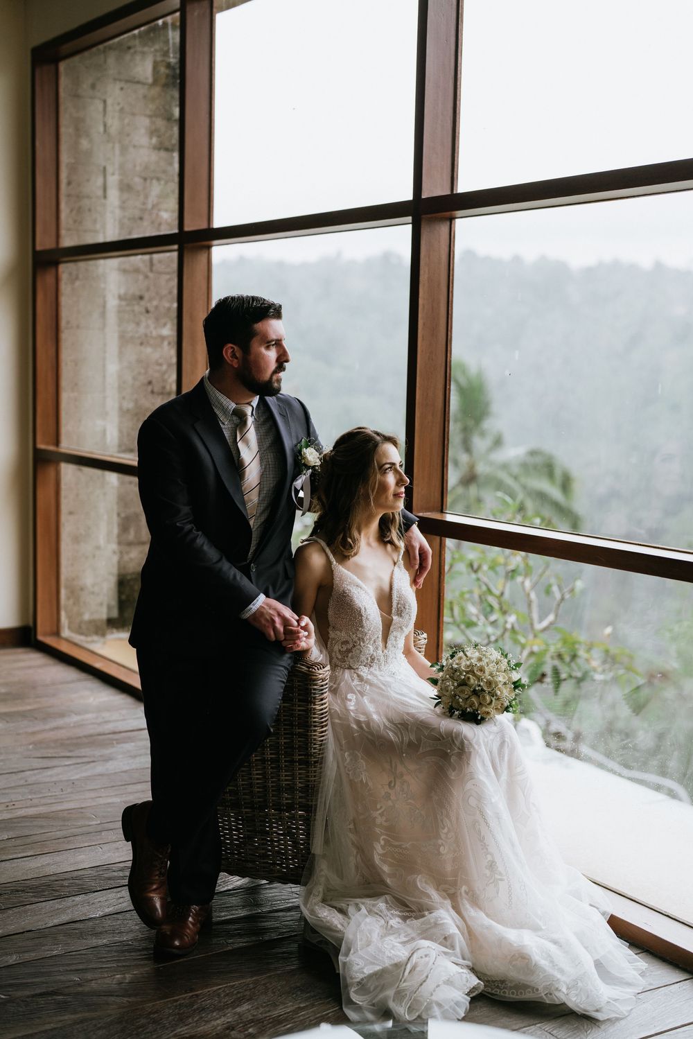 A couple in wedding attire sits together by large windows overlooking a scenic mountain landscape.