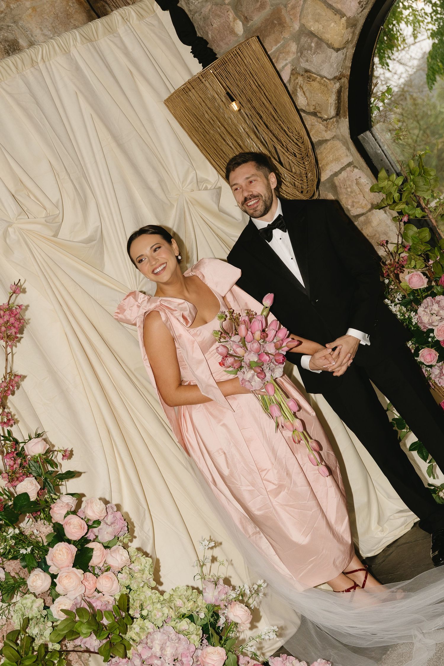 A couple poses together at their wedding among pink floral decorations and a stone wall backdrop.