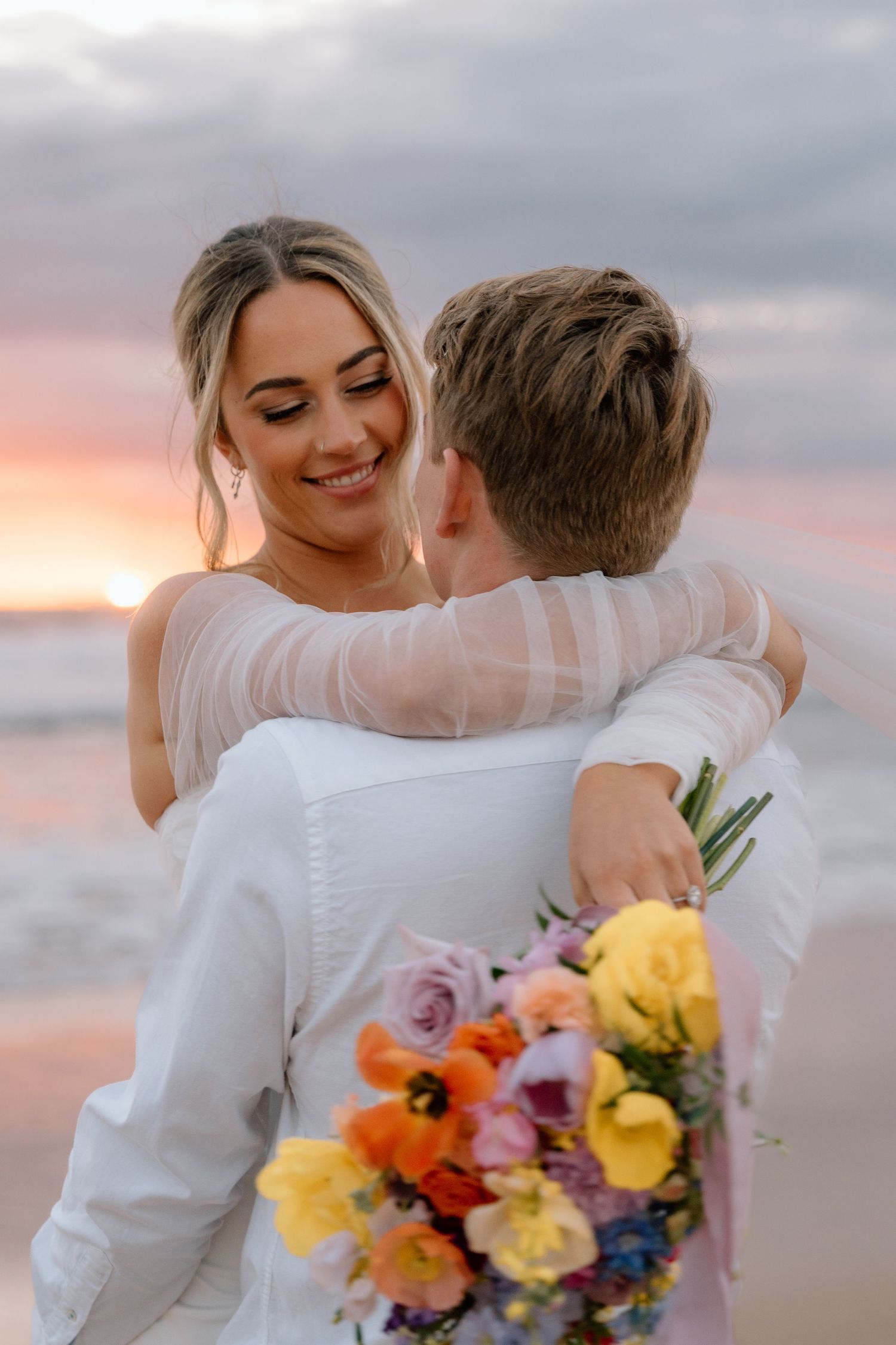 Romantic beach wedding couple embraces at sunset with colorful bouquet featuring yellow and purple flowers.