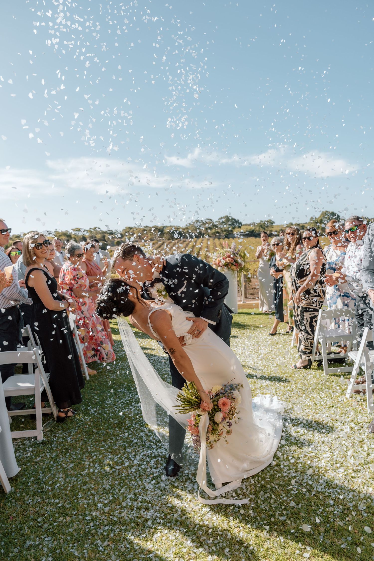 Confetti fills the air as wedding guests celebrate on a grassy lawn under bright blue skies.