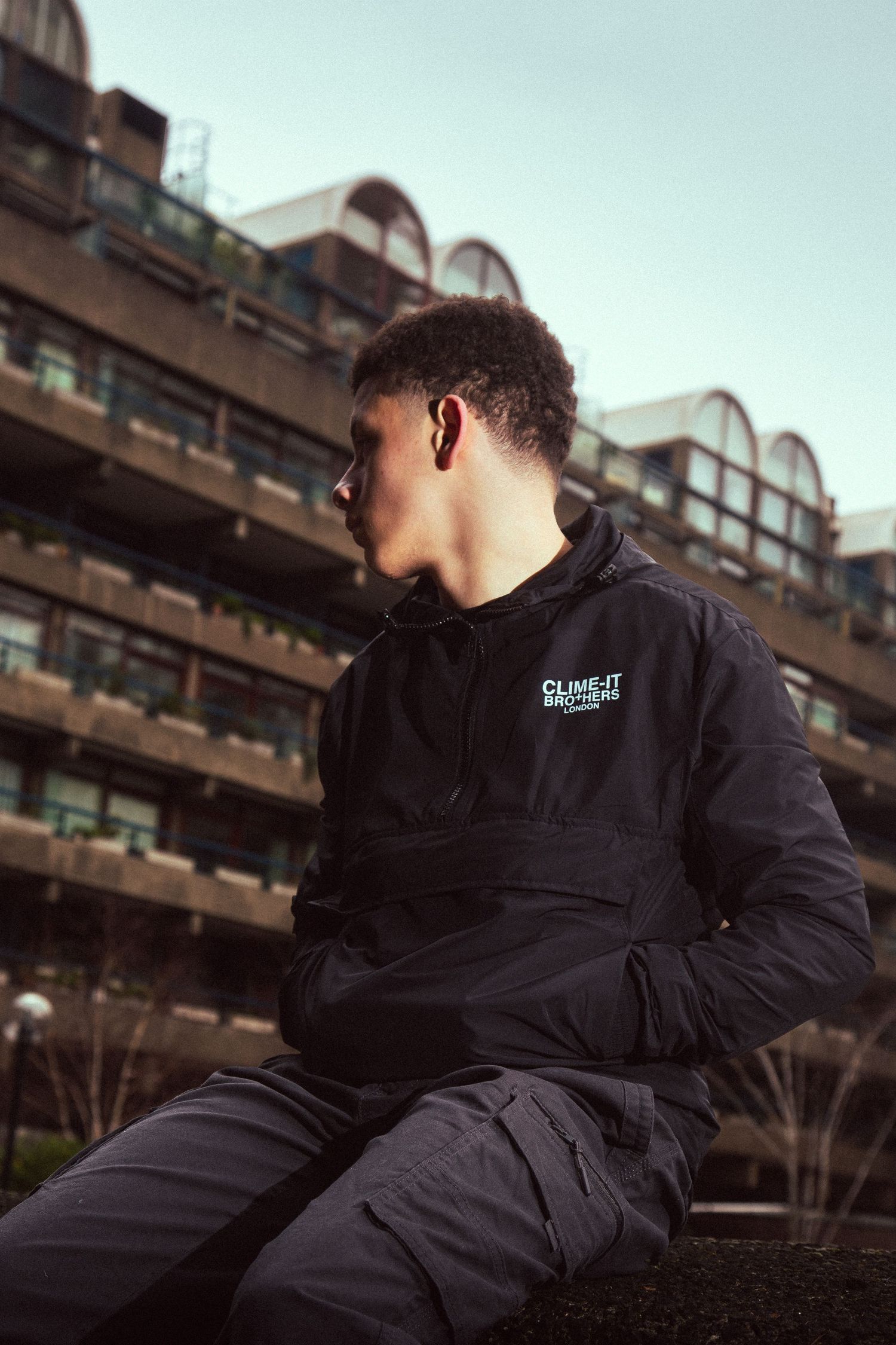Person wearing black jacket sits on concrete ledge against urban architecture in moody lighting.