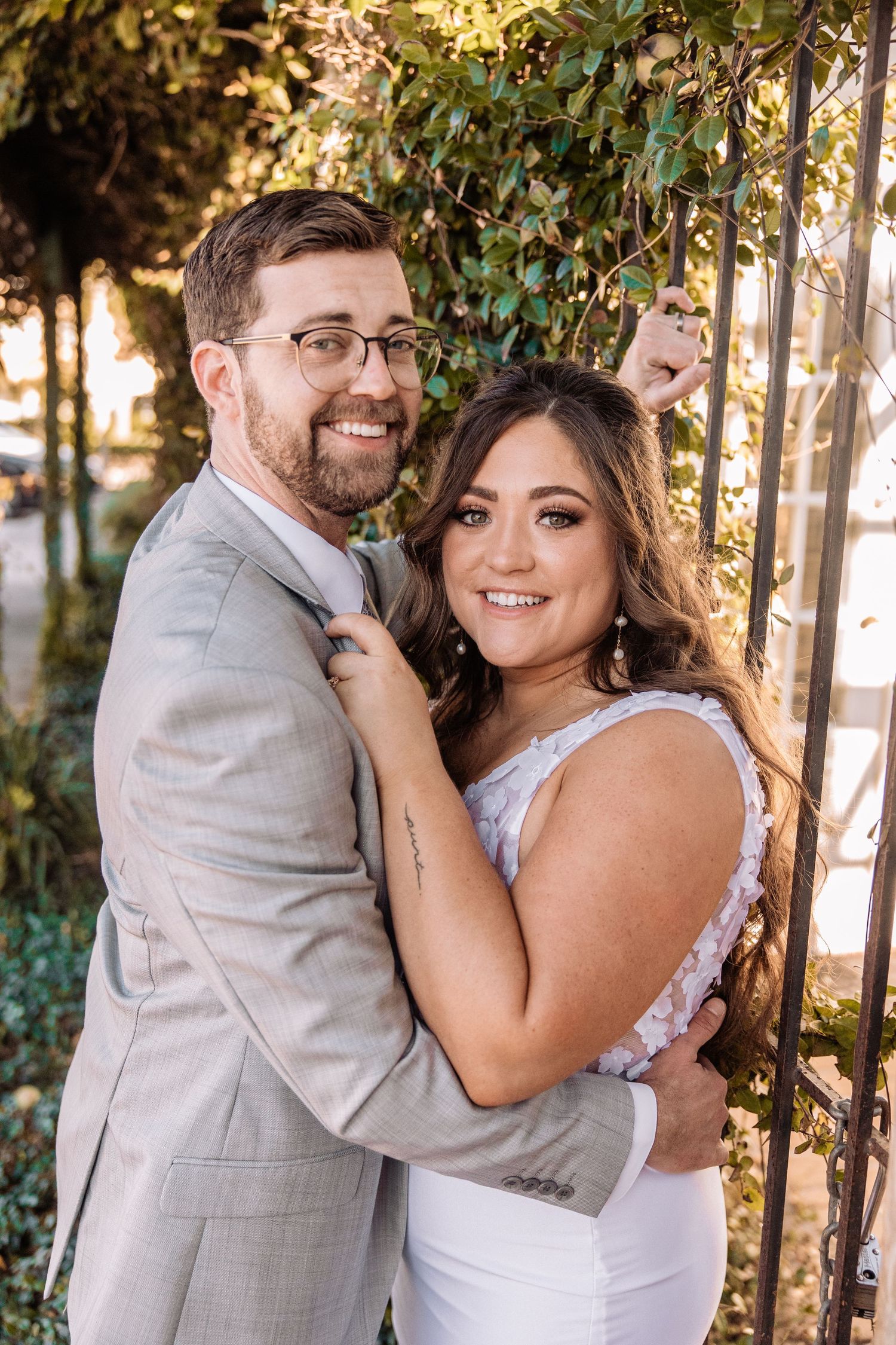 Joyful wedding couple shares laughs against sunlit greenery and rustic backdrop.