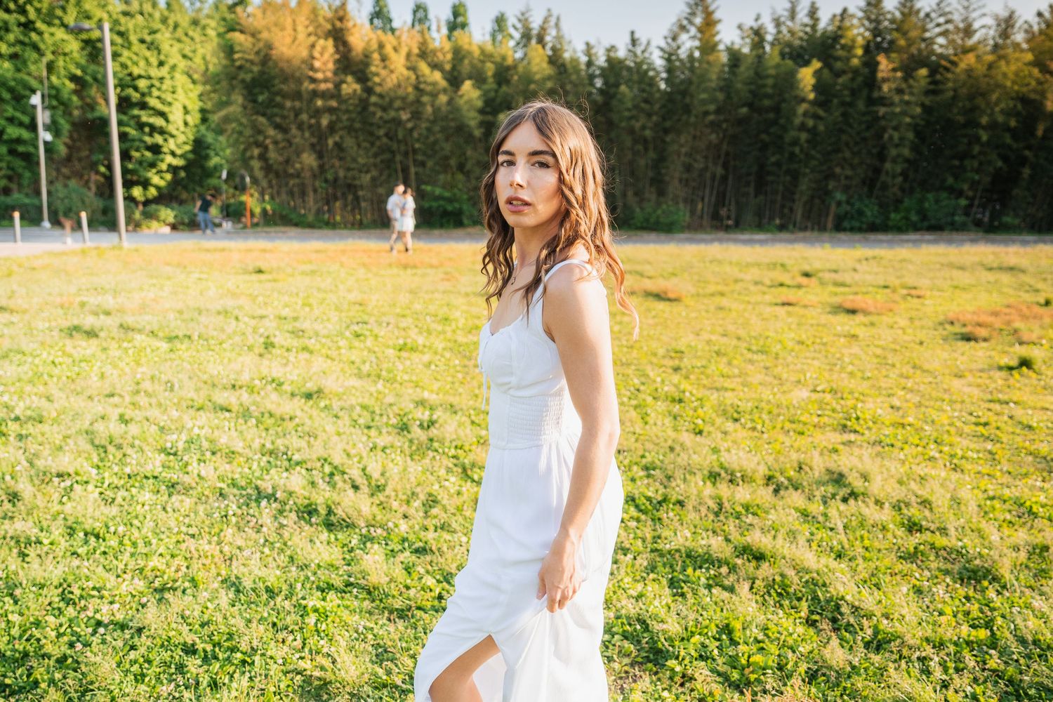 Person in a flowing white dress twirls joyfully in a sunlit meadow with evergreen trees in the background.